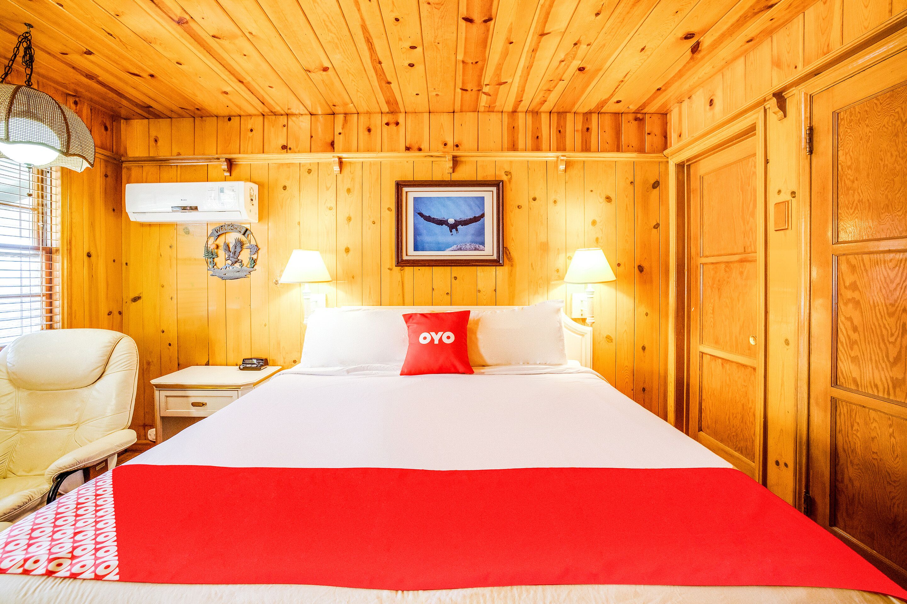 Una habitación con paneles de madera, una cama, dos lámparas de noche, una silla, un equipo de aire acondicionado y un cuadro con un águila.
