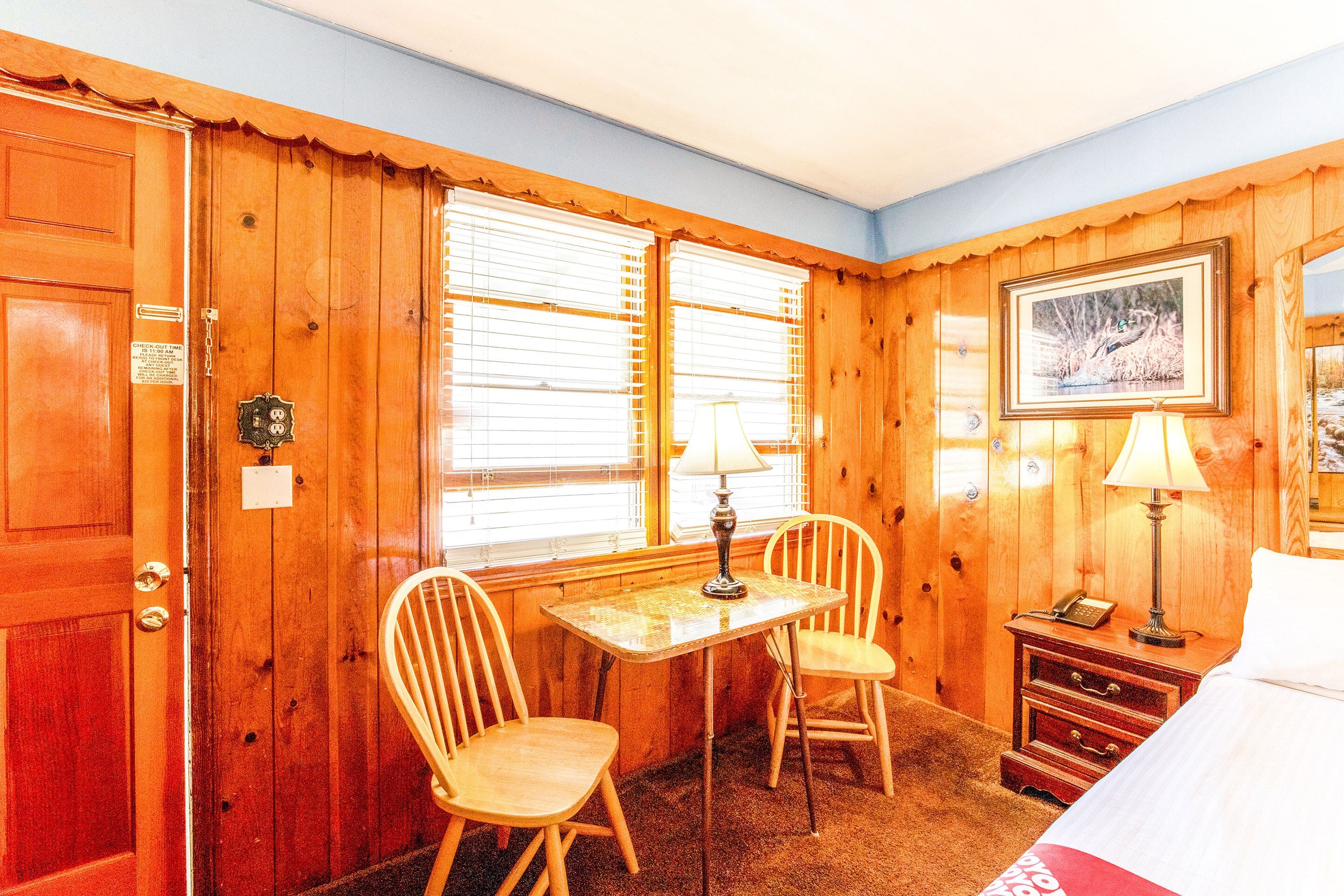 A wooden-paneled room with a bed, a table with chairs, a lamp, and a framed picture on the wall.