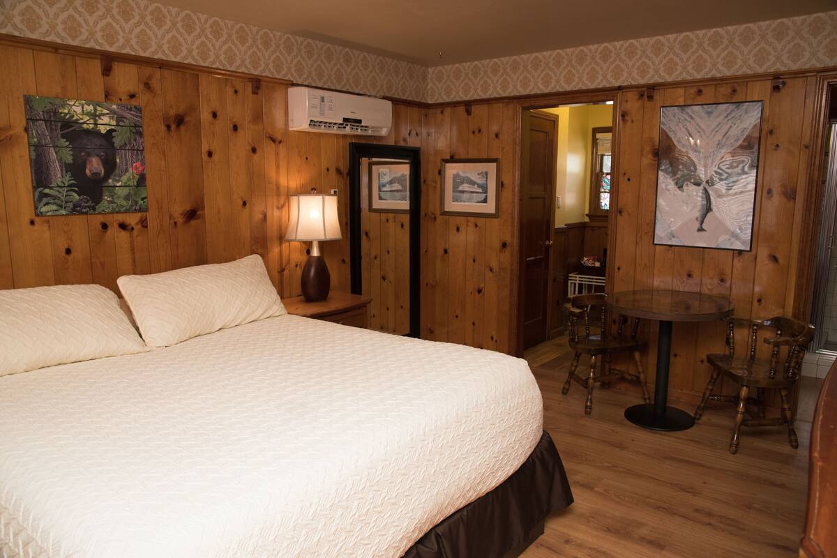 A bedroom with a large bed, a bedside table with a lamp, a wooden dining table with chairs, and framed pictures on the walls.