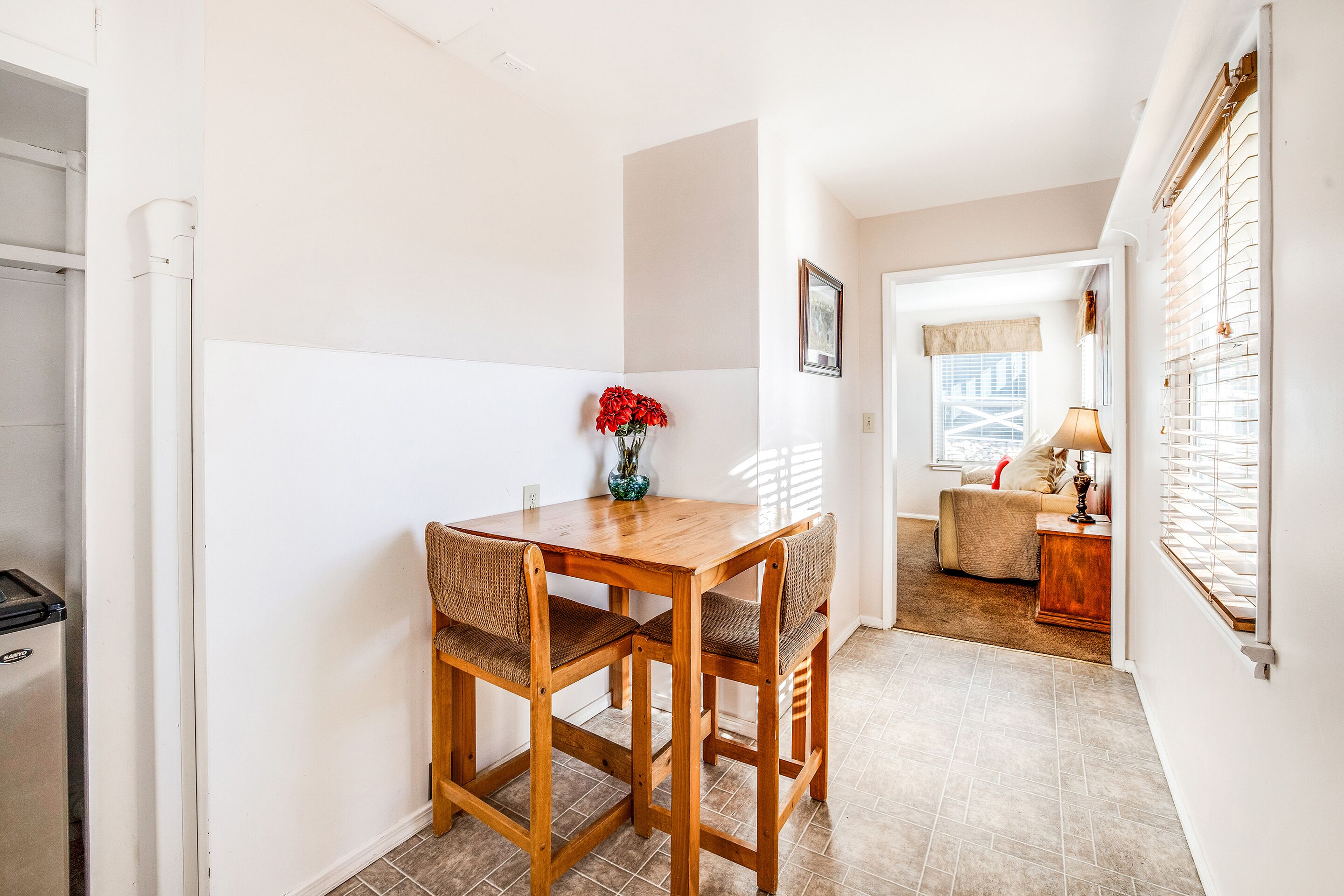 Uma área de jantar com mesa e cadeiras de madeira, um vaso com flores vermelhas e vista para uma sala de estar com sofá e luminária.