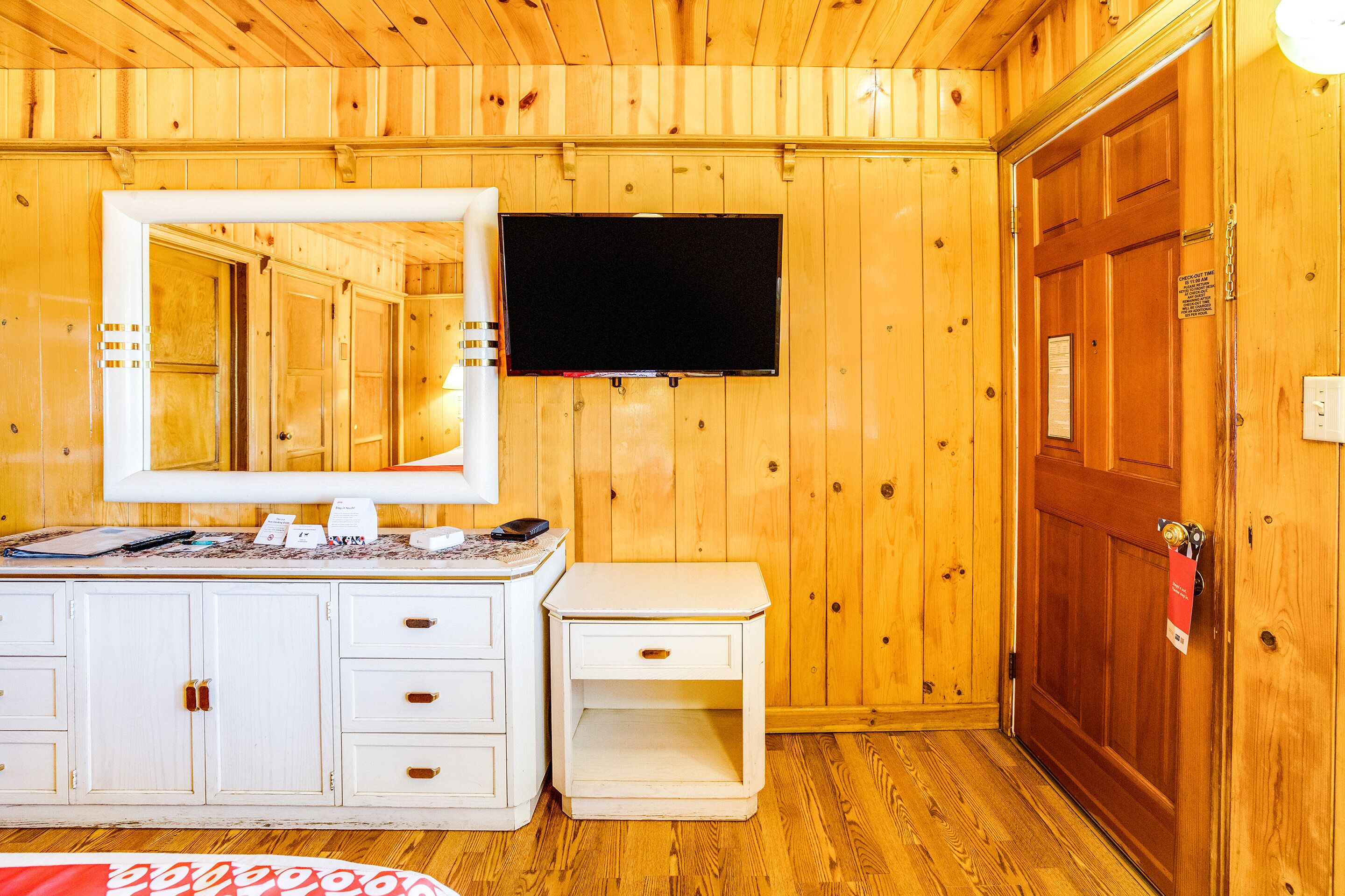 A wooden-paneled room with a white dresser, a mirror, a flat-screen TV, and a wooden door.