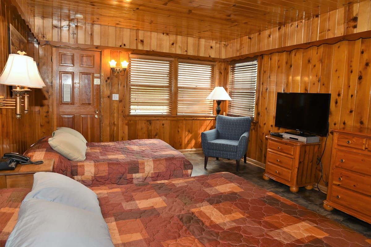 A wooden cabin room with two beds, a chair, a TV, and a dresser.