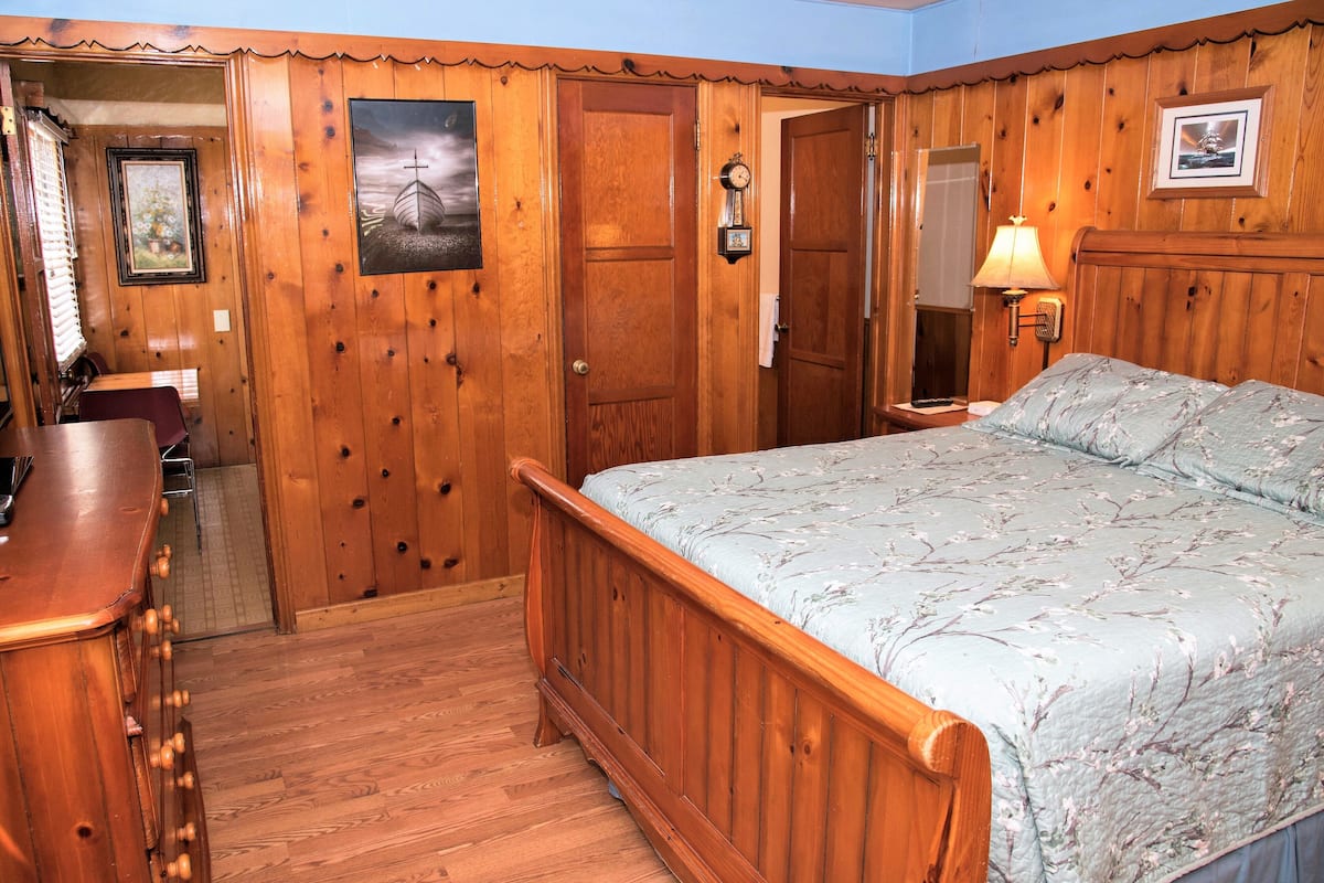 A wooden-paneled room with a bed, a desk, and a door leading to another room.