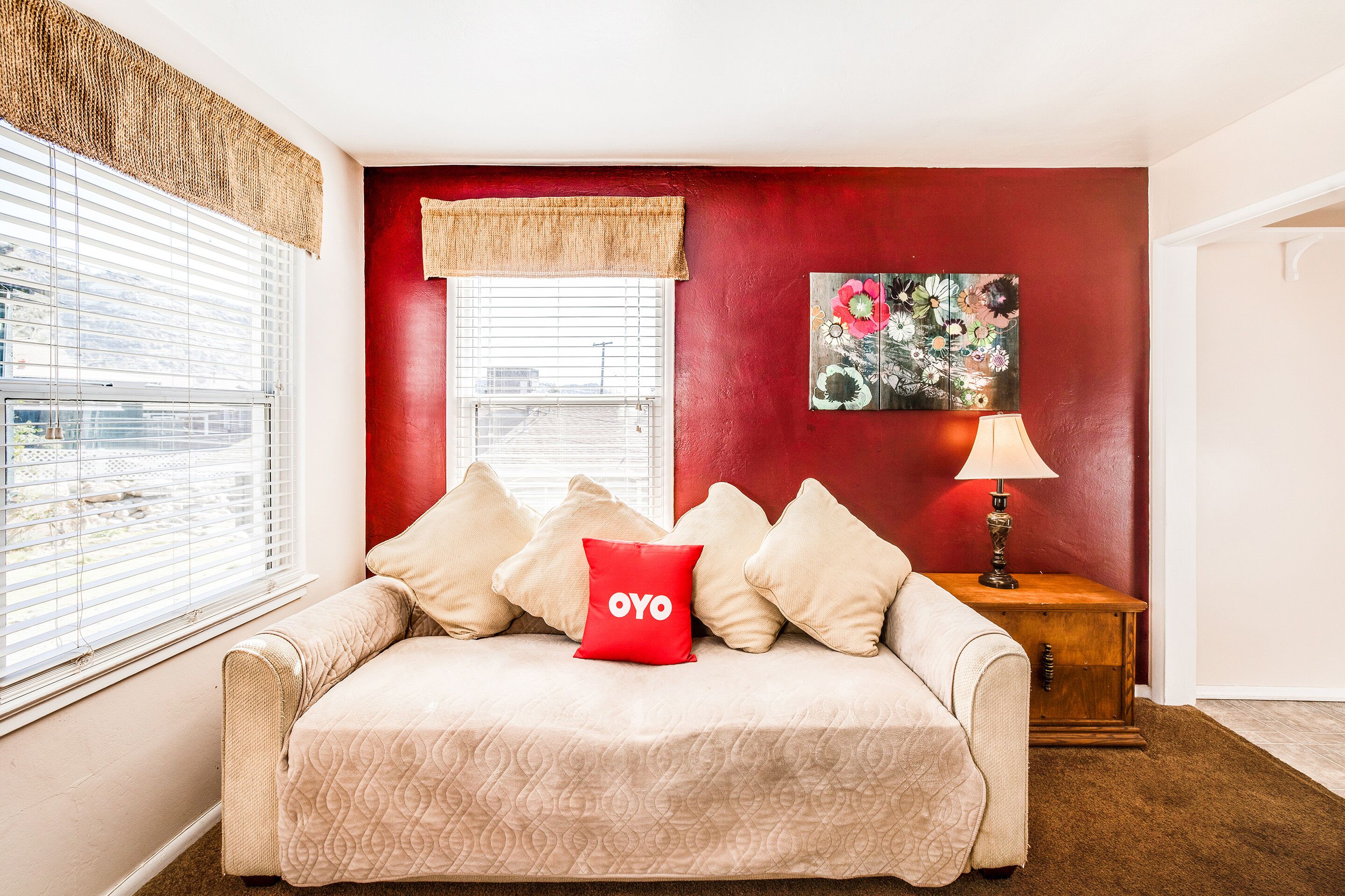 A cozy room with a beige sofa, a red accent wall, a window with blinds, a wooden side table with a lamp, and a floral painting.