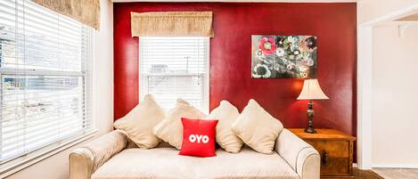 A cozy room with a beige sofa, a red accent wall, a window with blinds, a wooden side table with a lamp, and a floral painting.