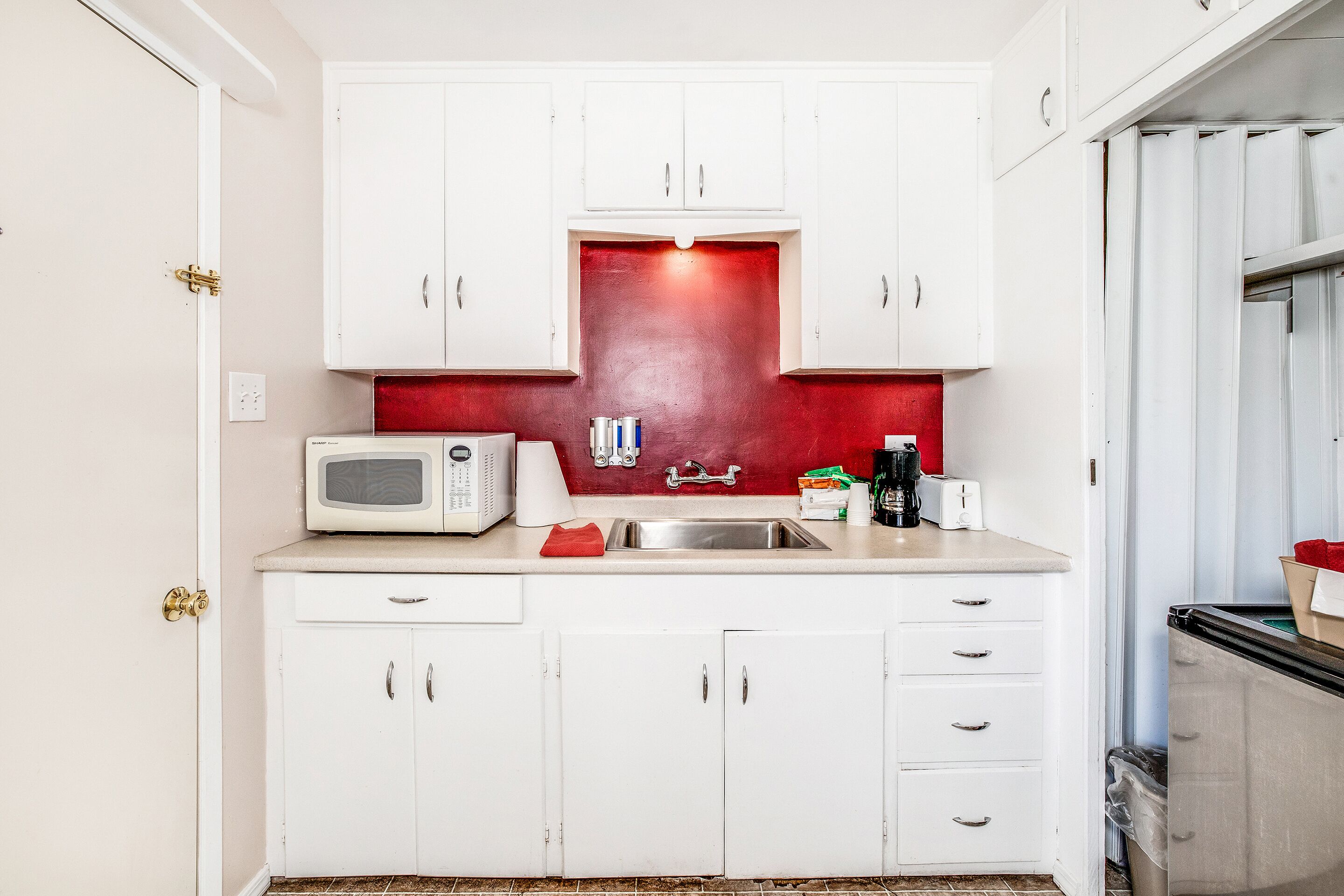 Una cucina compatta con mobili bianchi, uno schienale rosso, un forno a microonde, un lavandino e un cestino.