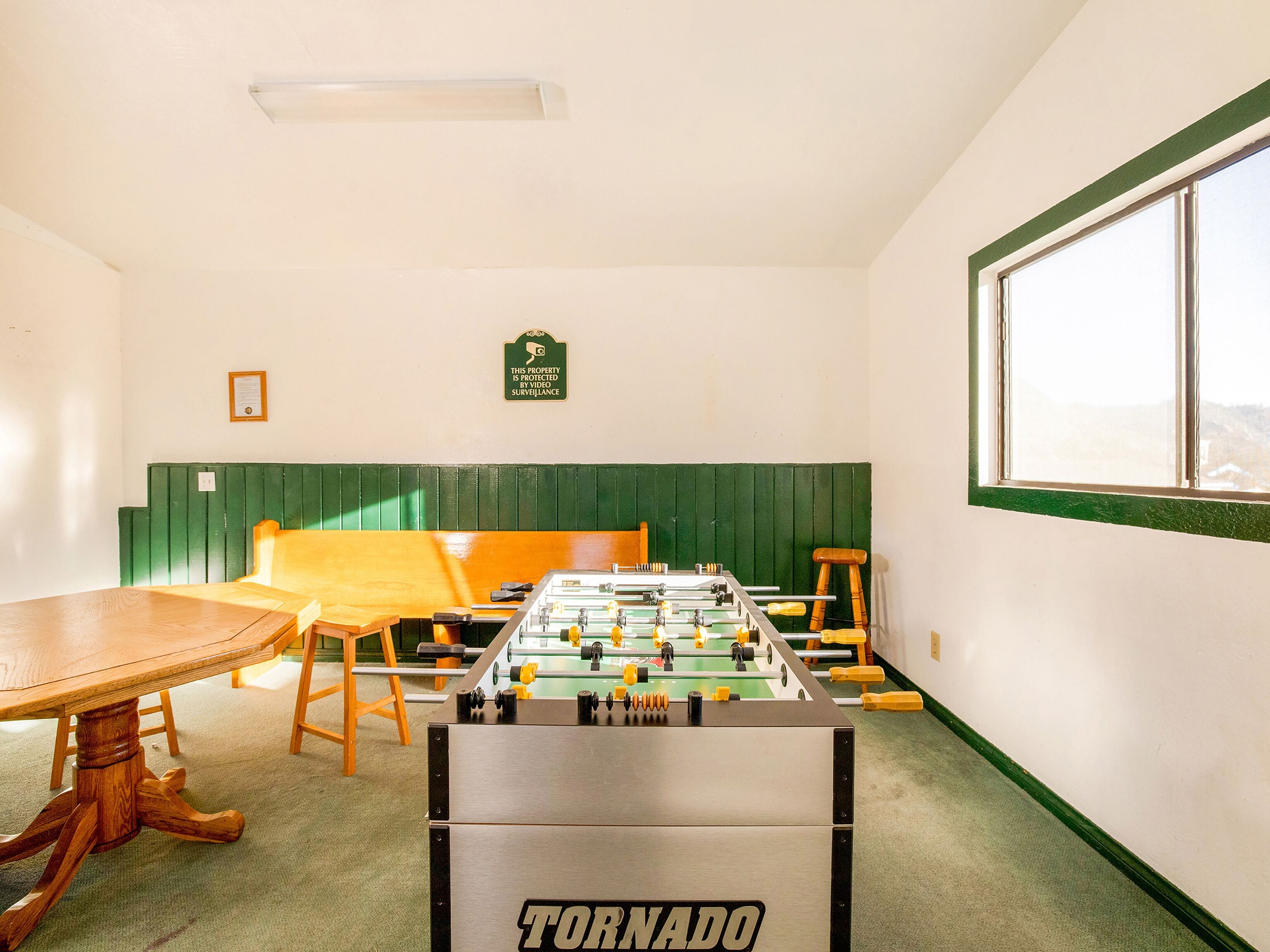 A room with a foosball table, a wooden table, and a window.