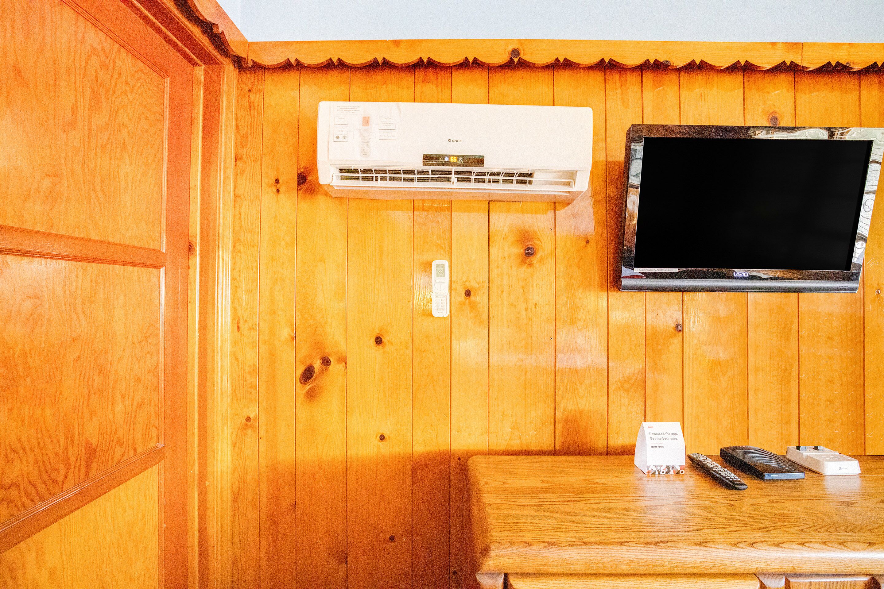 A wooden-paneled room with a mounted air conditioner, a television, and a table with remote controls and a power strip.