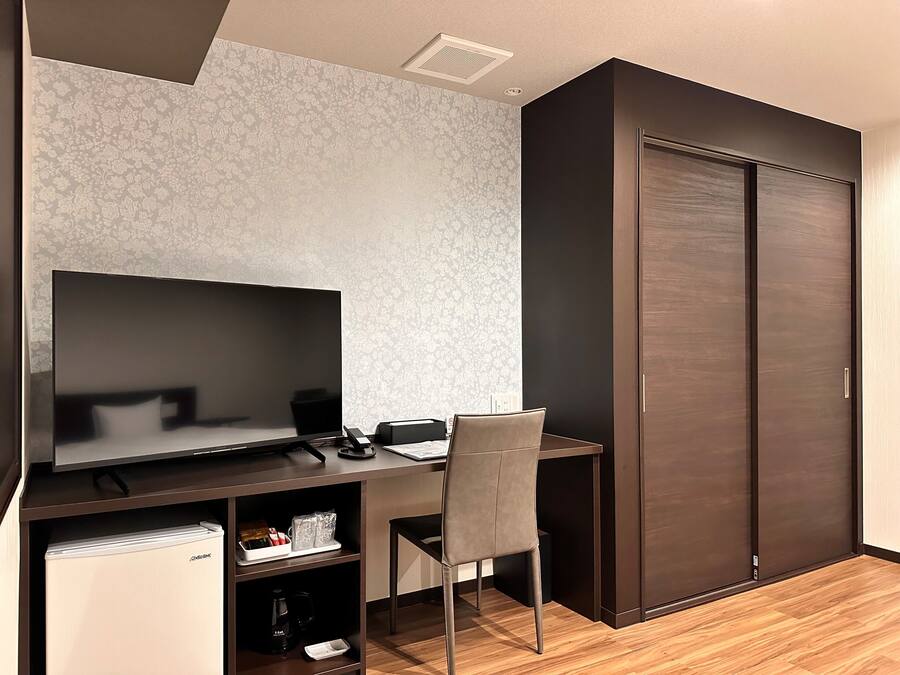 Una habitación moderna con un escritorio de madera oscura, un televisor de pantalla plana, una silla, un refrigerador pequeño y un armario.