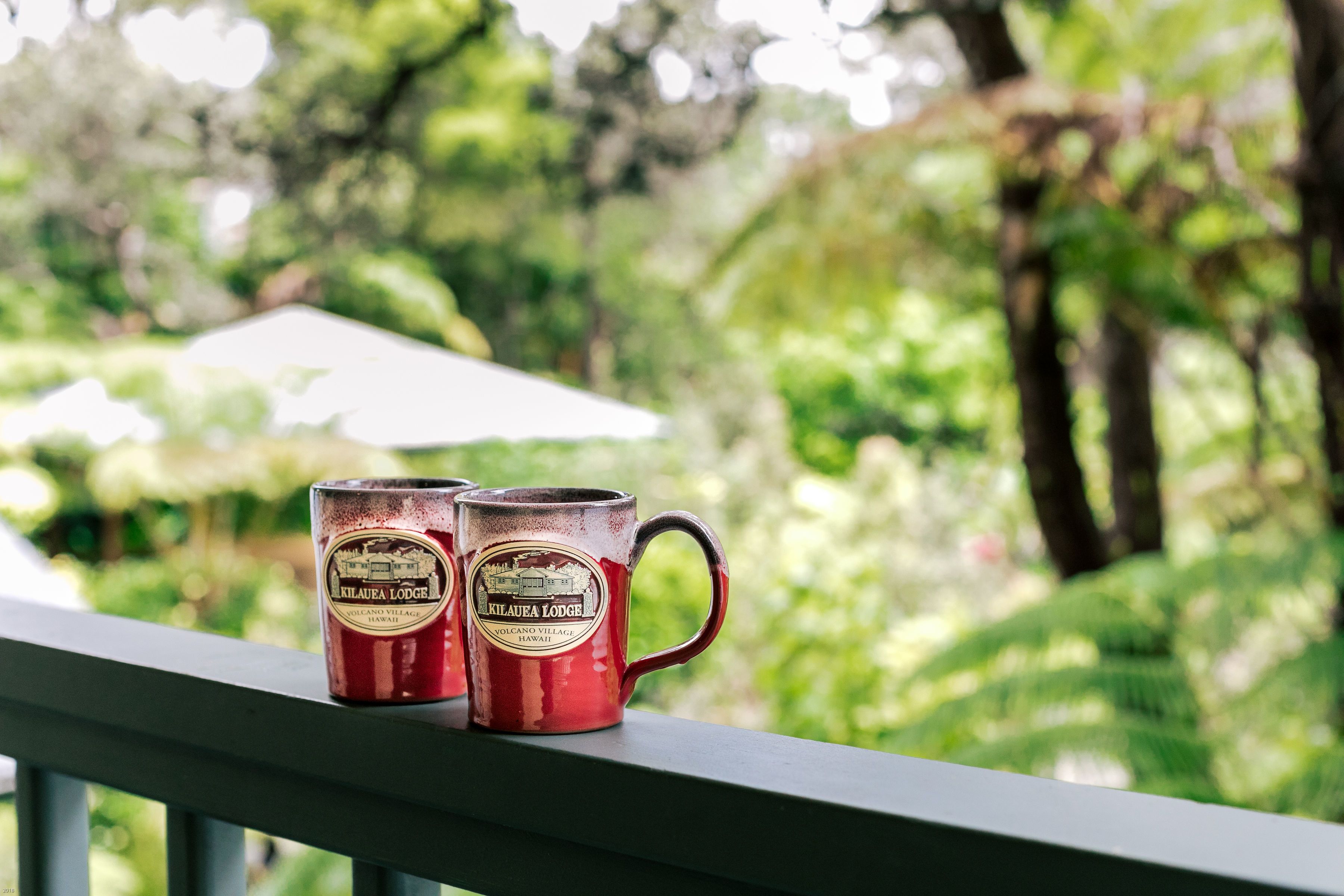 Dos tazas rojas con el texto 'Kilauea Lodge' sobre una barandilla de balcón, con un fondo verde desenfocado.