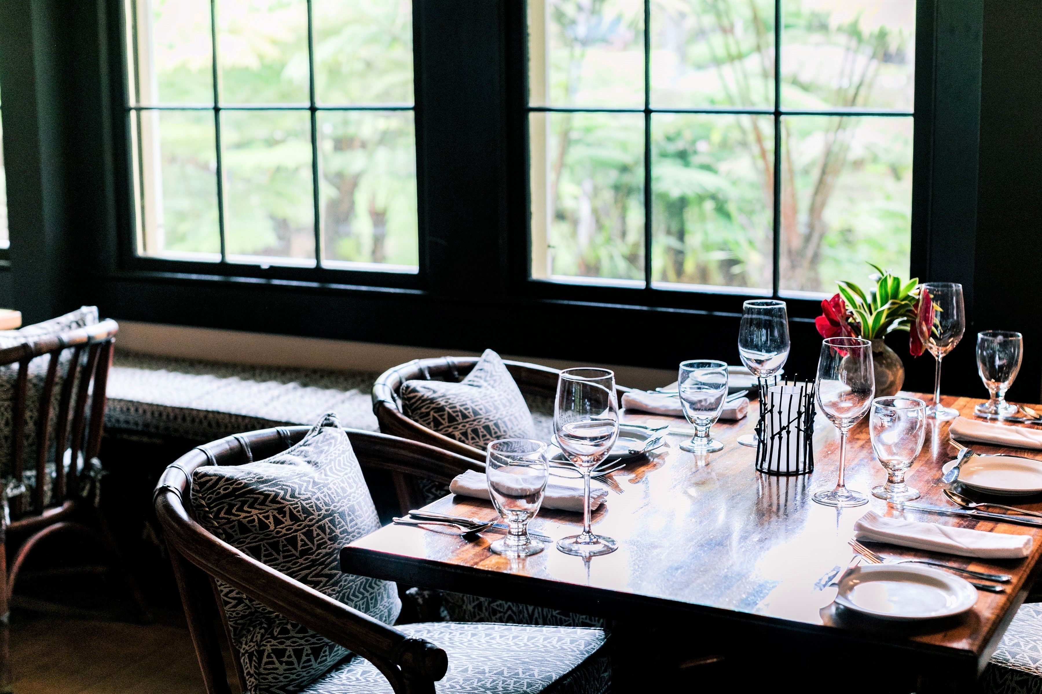 A dining table set with glassware and plates, surrounded by patterned chairs, with a view of greenery through large windows.