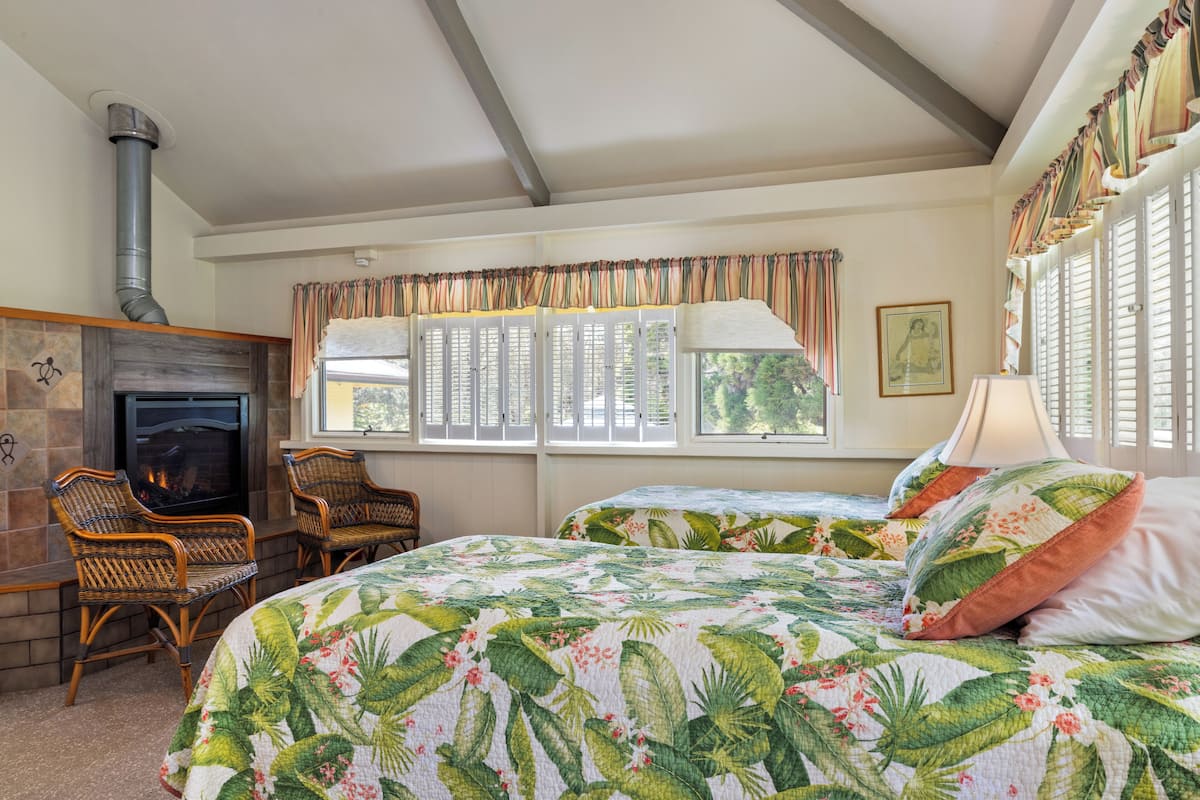 A cozy room with a fireplace, wicker chairs, a bed with a tropical print bedspread, and a large window with shutters.