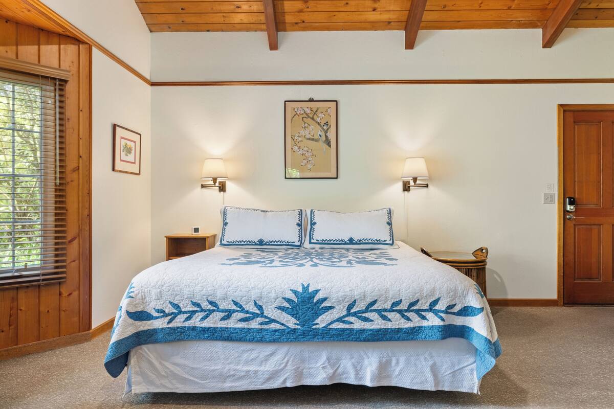 A bedroom with a large bed, wooden ceiling beams, two wall-mounted lamps, a framed picture, and a door.