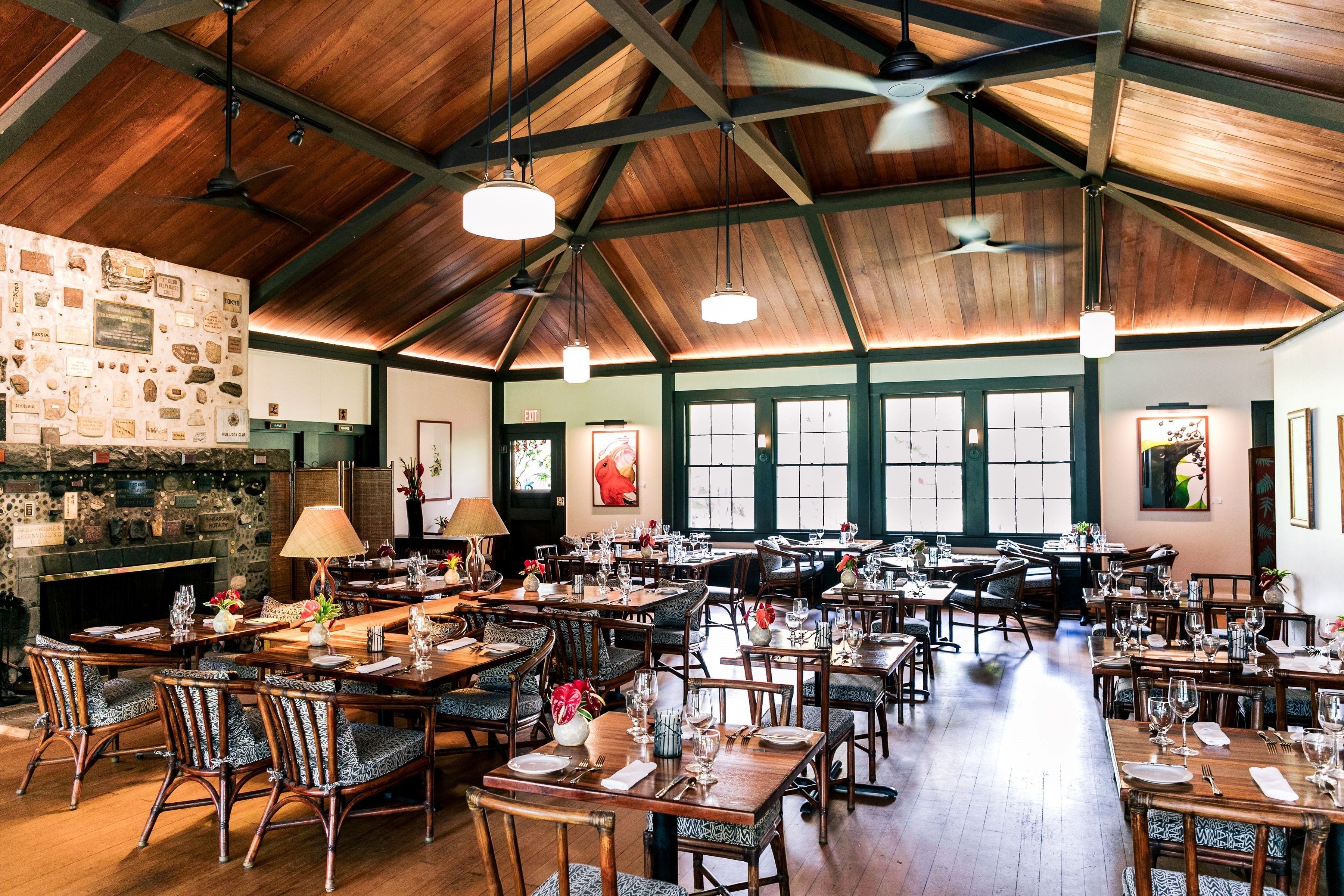 A spacious restaurant with wooden ceilings and walls, featuring multiple dining tables and chairs, a stone fireplace, and large windows.
