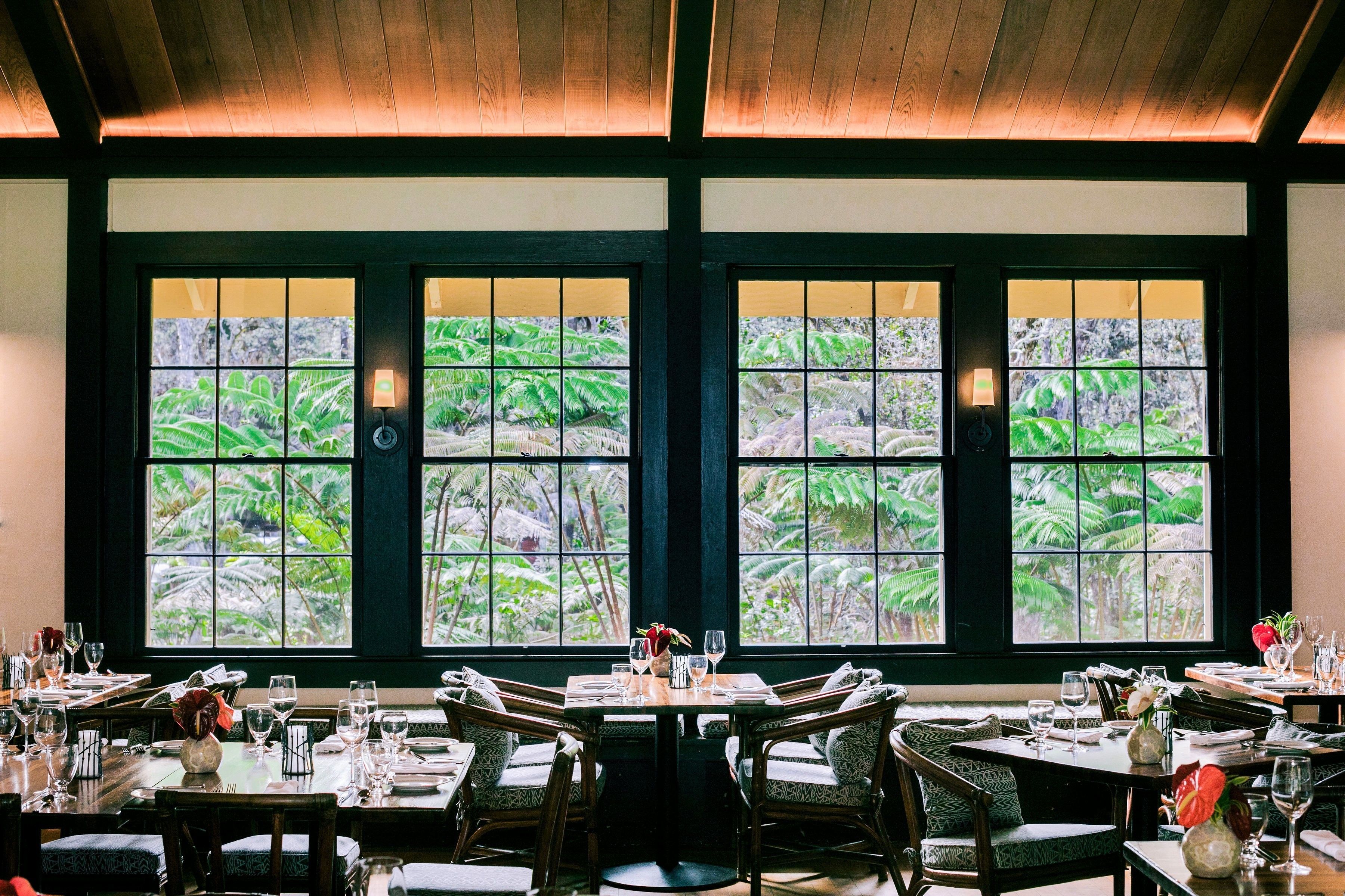 A restaurant with large windows offering views of lush greenery. Tables are set with wine glasses and plates.