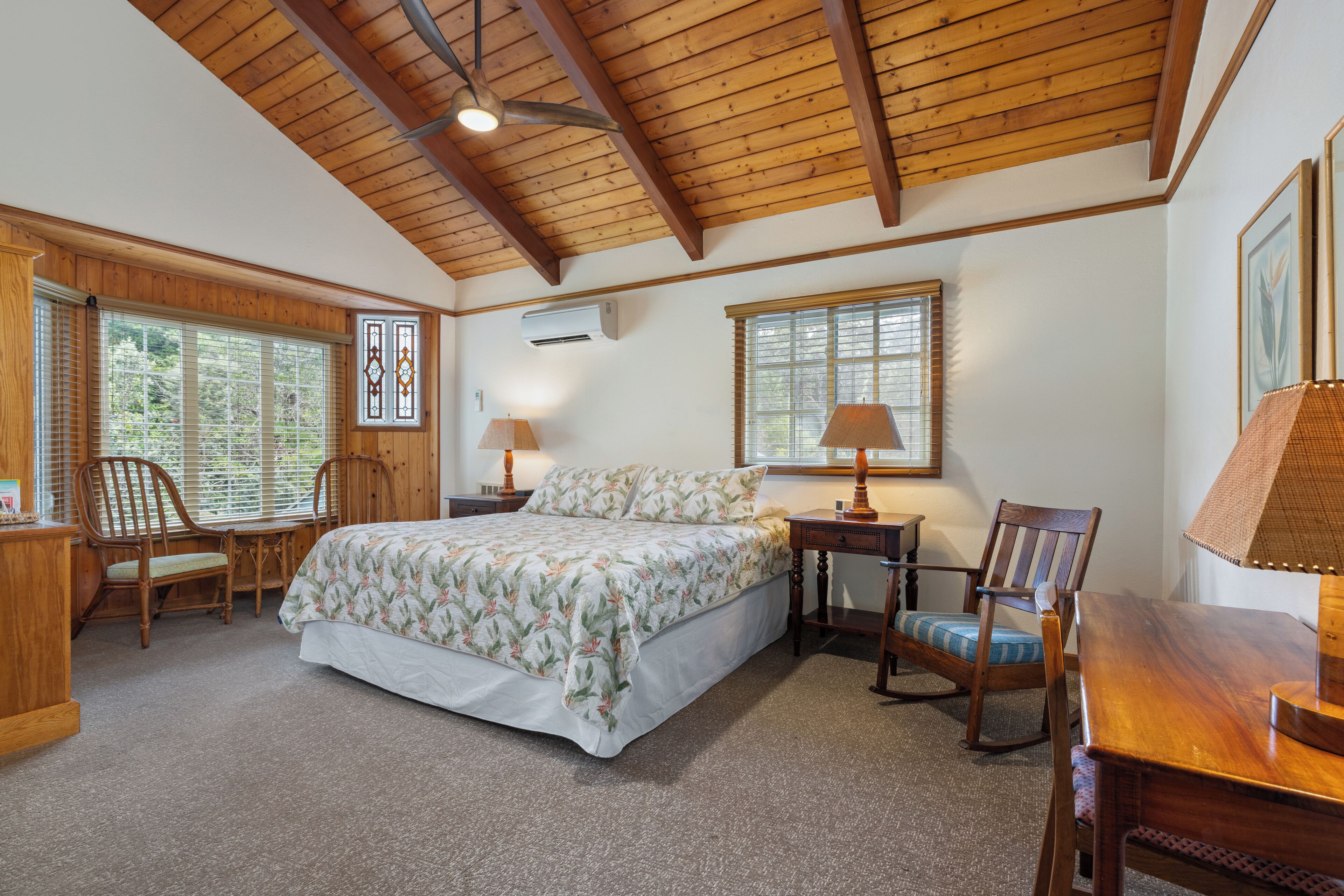 A bedroom with a wooden ceiling, a bed with floral bedding, two chairs, a table, and a window with blinds.