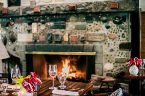Une table à manger dressée avec des verres à vin et un vase de fleurs, une cheminée où brûle un feu, et un mur de pierre sur lequel sont exposés divers objets.