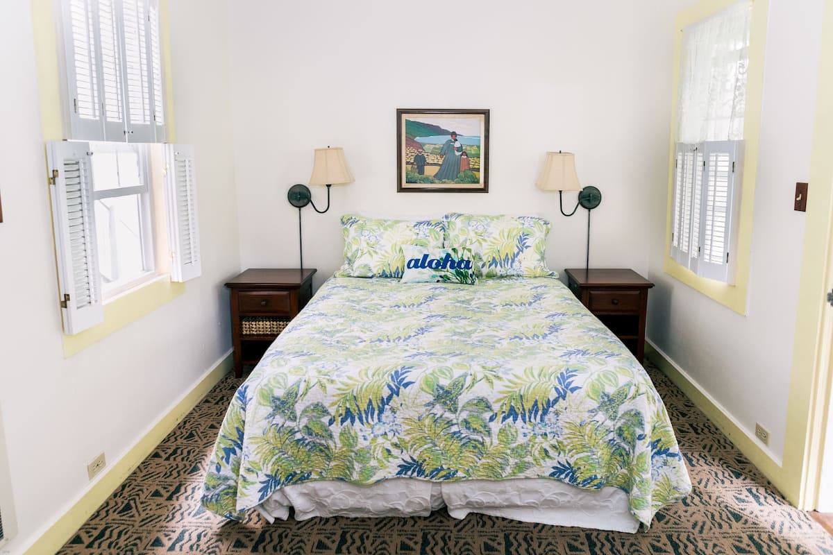 A bedroom with a bed, two nightstands, two wall-mounted lamps, a framed picture, and a window with shutters.