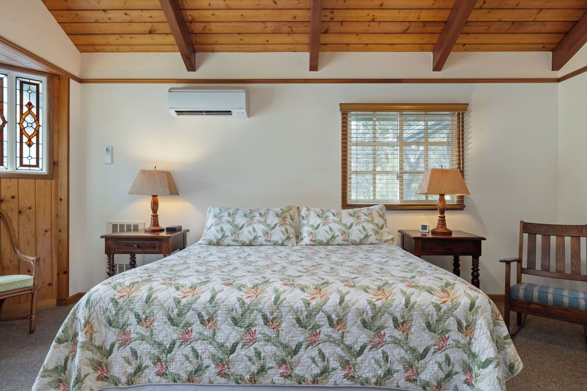 A bedroom with a large bed, two bedside tables with lamps, a wooden chair, a window with blinds, and an air conditioning unit on the wall.