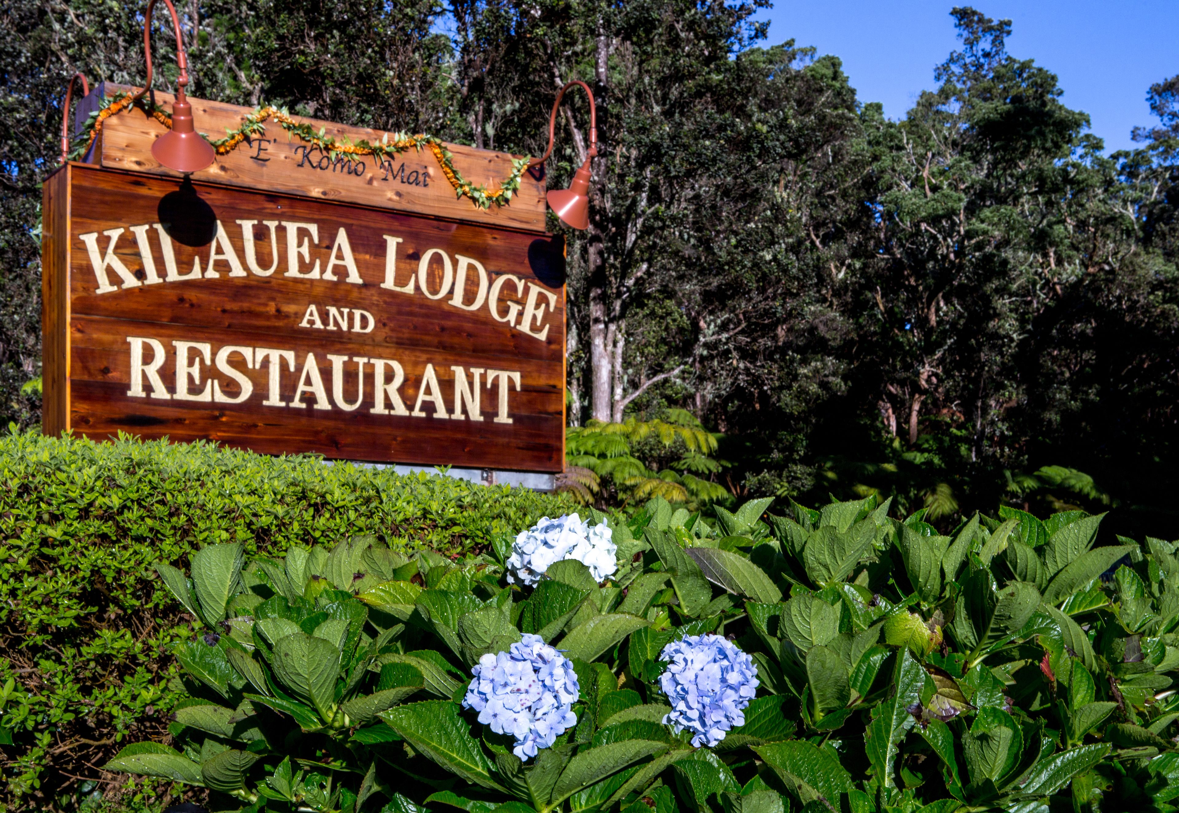 Un letrero de madera para 'Kilauea Lodge and Restaurant' con un cascabel y una guirnalda decorativa.