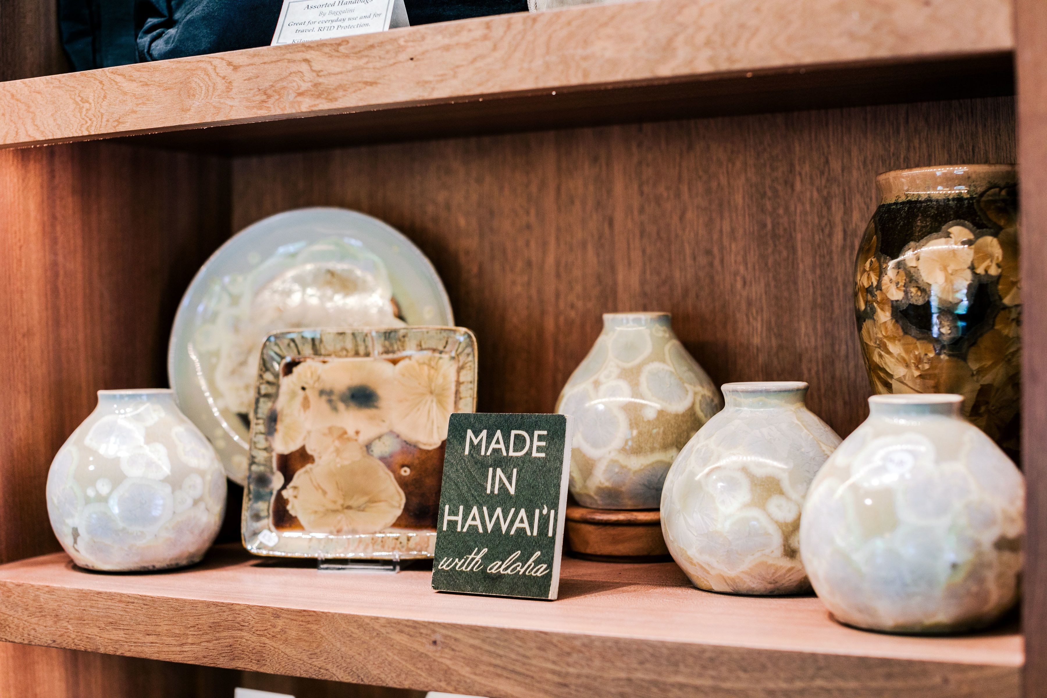Una repisa de madera exhibe varios jarrones de cerámica con un letrero que indica que fueron hechos en Hawaii con cariño.
