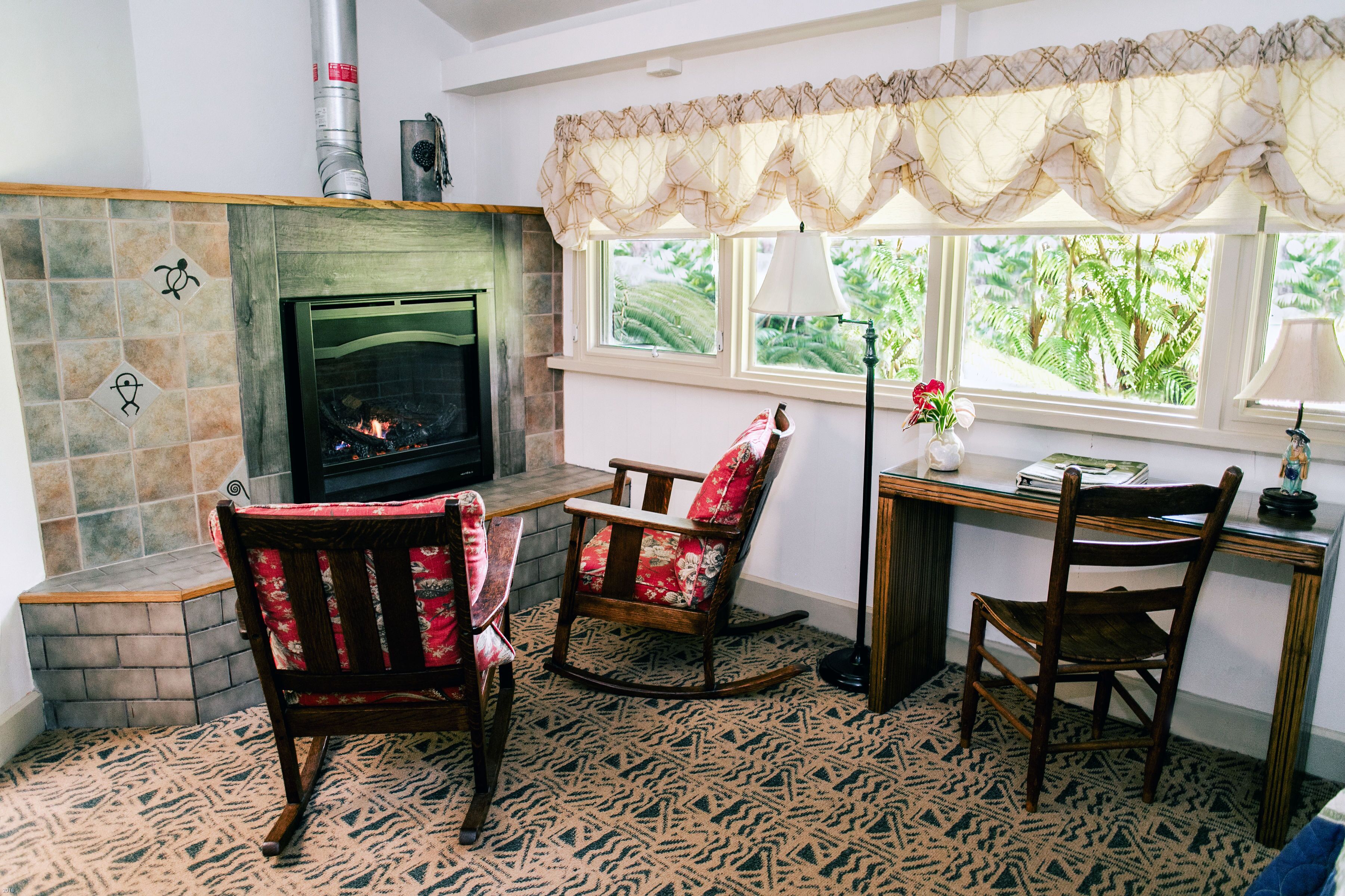 A cozy living room with a fireplace, two rocking chairs, a desk with a lamp, and a view of greenery outside.