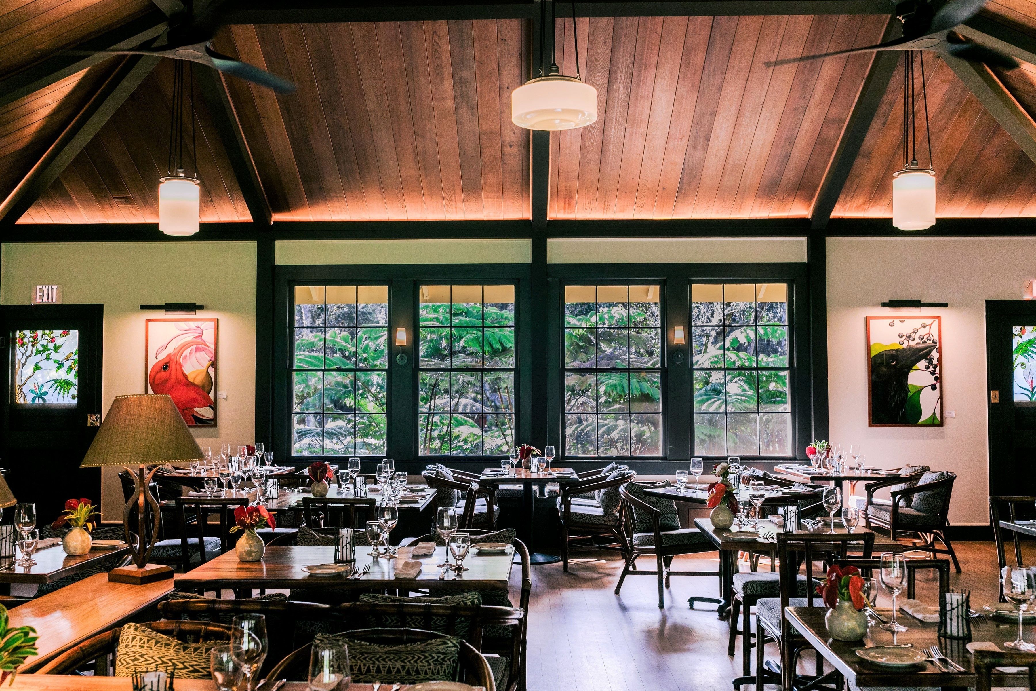 A restaurant with multiple dining tables and chairs, large windows with a view of greenery, and a wooden ceiling.