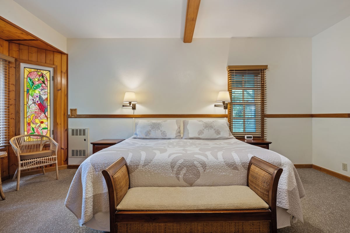 A bedroom with a large bed, two bedside lamps, a chair, a stained glass window, and a window with blinds.