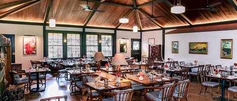 A spacious dining area with wooden tables and chairs, large windows, and ceiling fans.