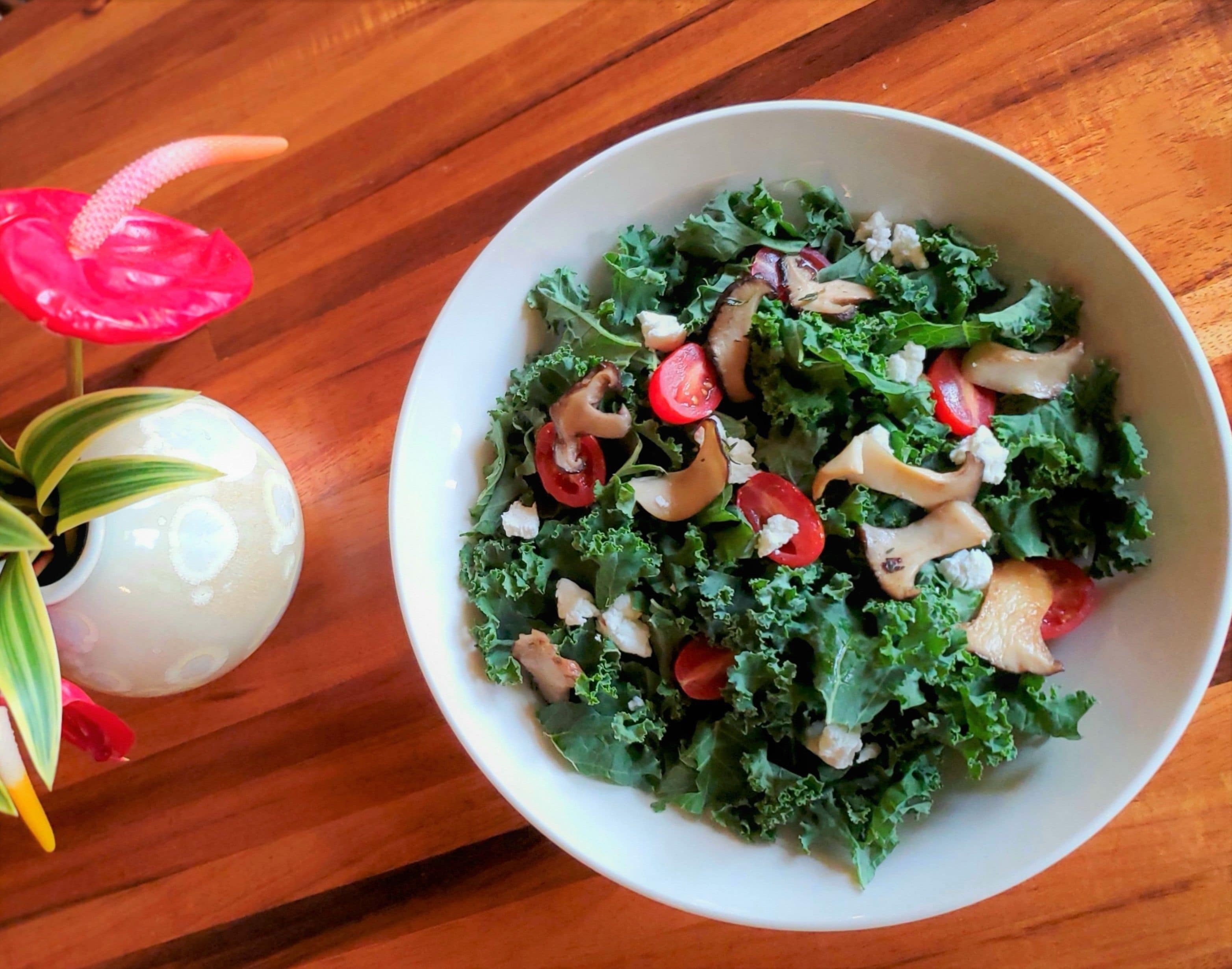 A bowl of salad with kale, cherry tomatoes, and mushrooms on a wooden table.