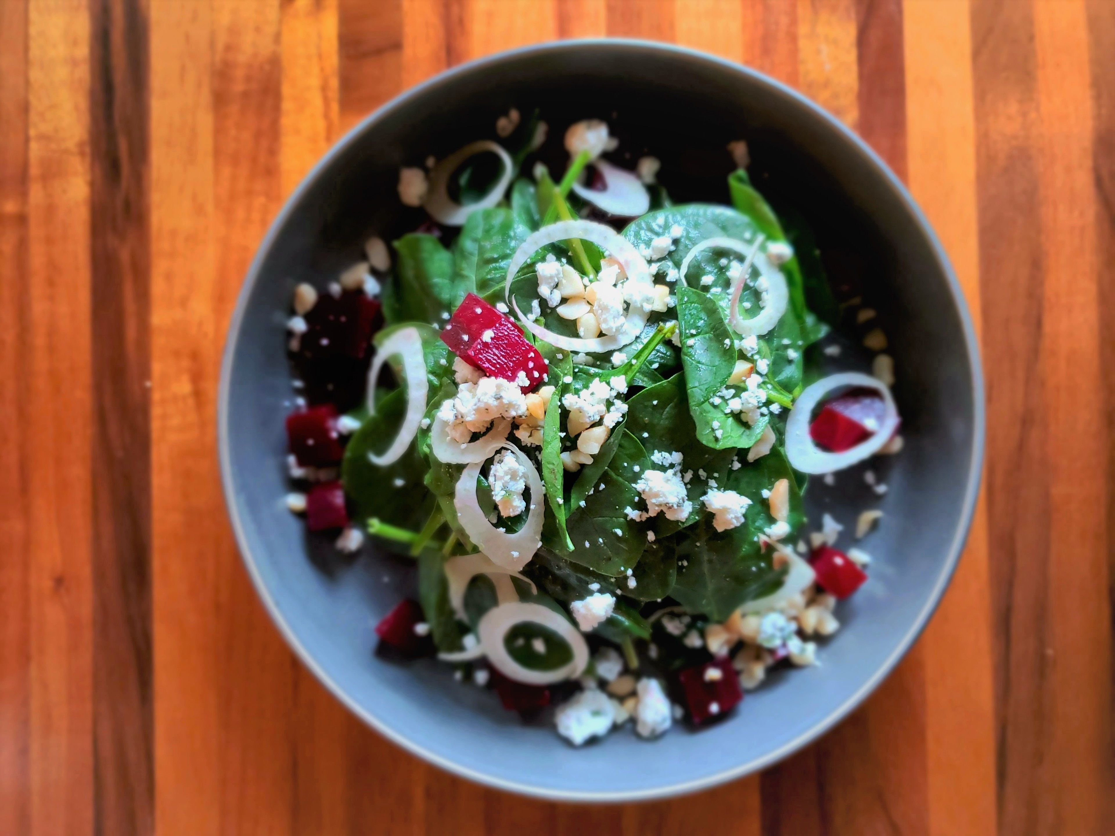 A bowl of salad with spinach, red beets, onions, and crumbled cheese on a wooden table.