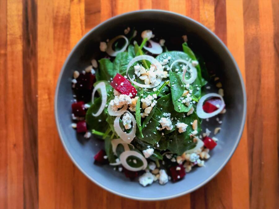 A bowl of salad with spinach, red beets, onions, and crumbled cheese on a wooden table.