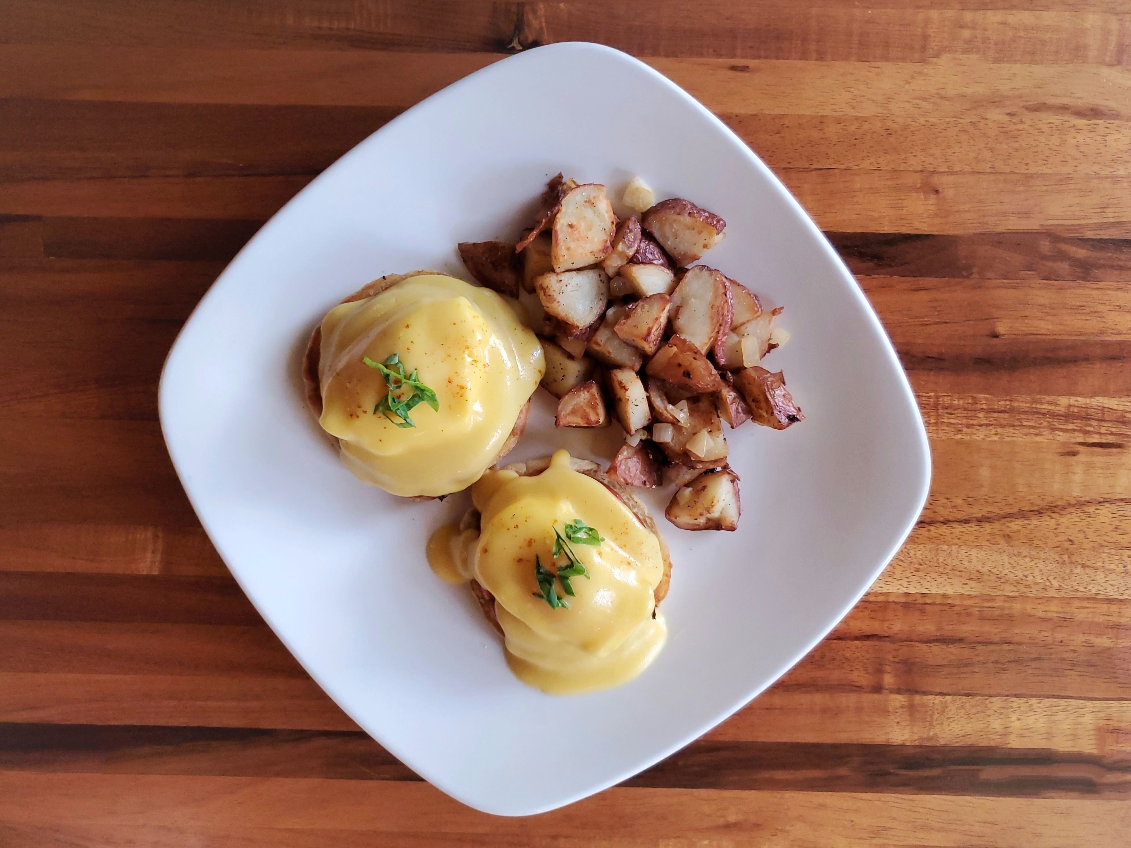 A white plate with two eggs benedict and roasted potatoes on a wooden table.