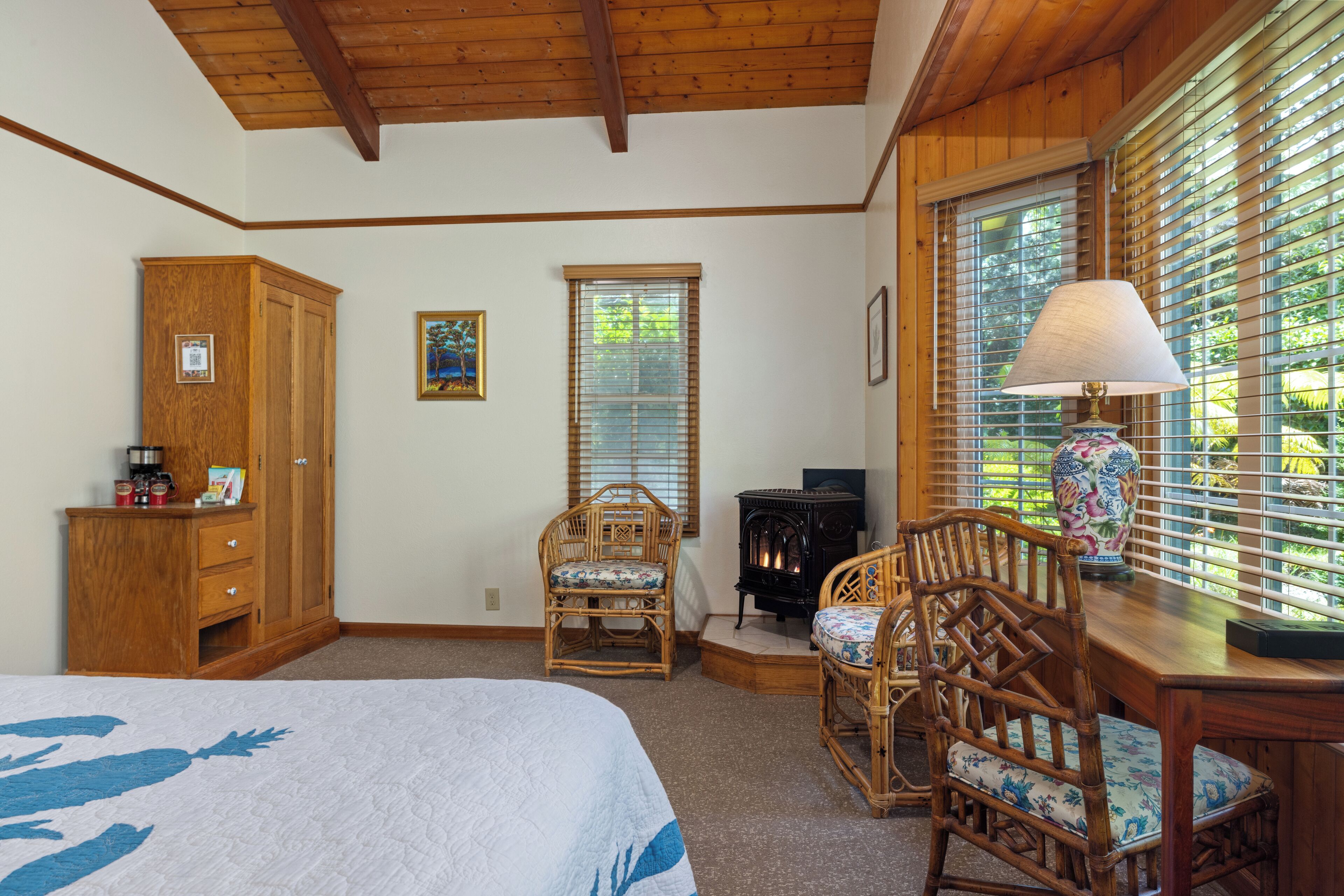 A bedroom with a bed, wooden wardrobe, two wicker chairs, a table with a lamp, and a fireplace.