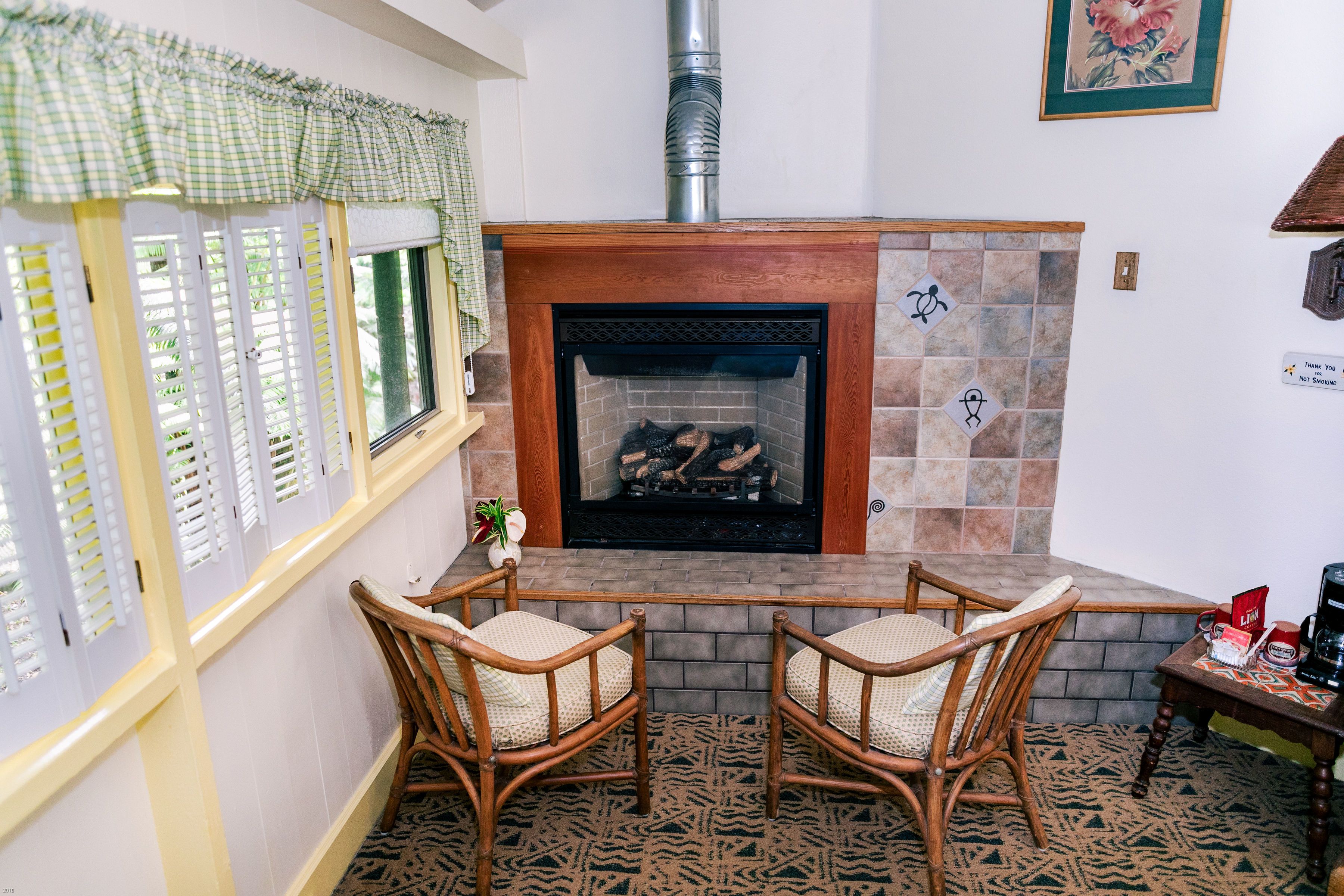 Una habitación acogedora con chimenea, dos sillones de mimbre, una mesita y una ventana con contraventanas.