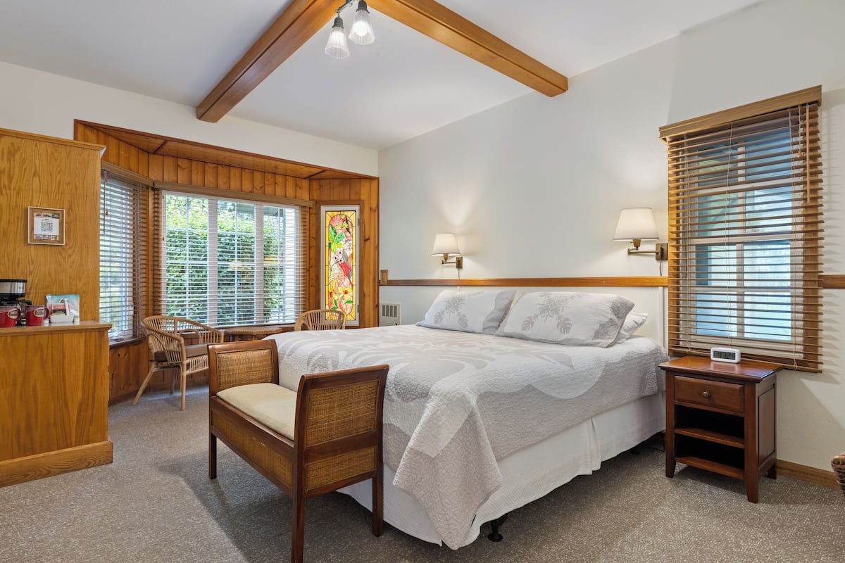 A bedroom with a large bed, wooden headboard, two bedside tables with lamps, a chair, a desk with a computer, and a stained glass window.
