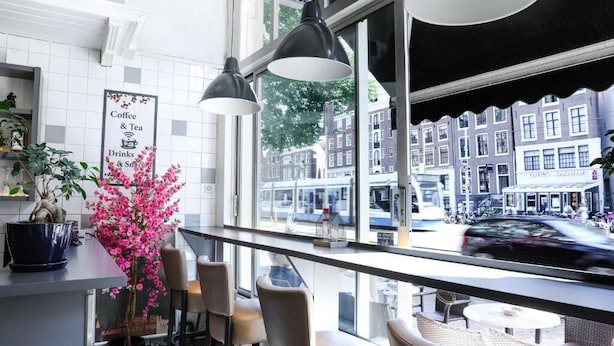 A café with a view of a street, a sign indicating coffee, tea, drinks, and snacks, and a potted plant with pink flowers.