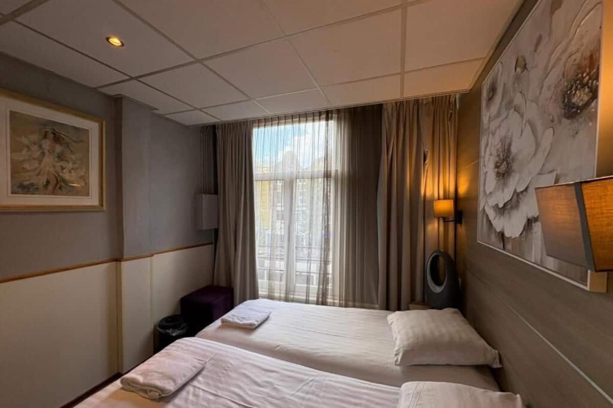 A hotel room with two beds, a large window with curtains, a wall-mounted lamp, and a framed artwork.