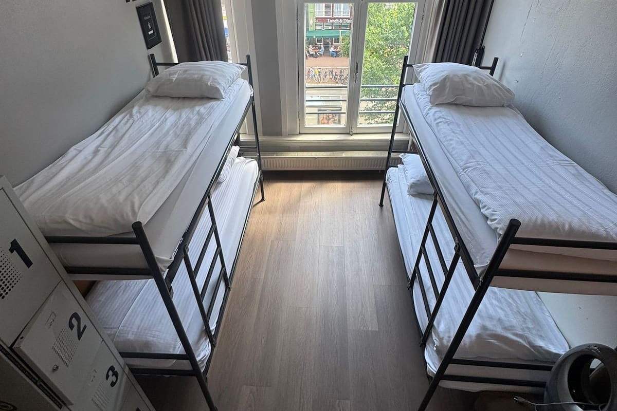A dormitory room with two single beds, a window with a view of buildings, and a ceiling with a smoke detector.