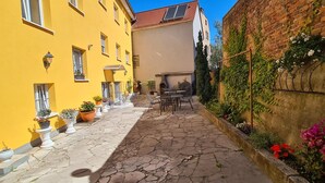 Un estrecho callejón adoquinado, con un edificio amarillo a un lado y un muro de ladrillo al otro, decorado con plantas en macetas y mesas al aire libre.