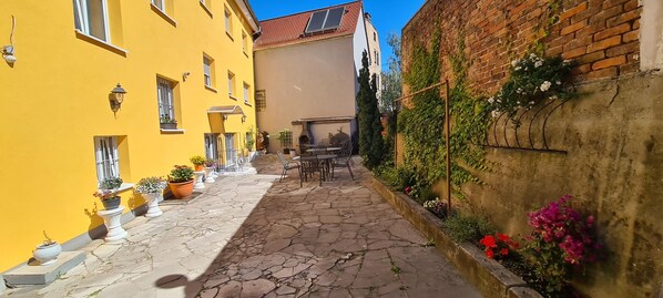 Une ruelle étroite pavée, avec un bâtiment jaune d’un côté et un mur de briques de l’autre, agrémentée de plantes en pot et de sièges en plein air.