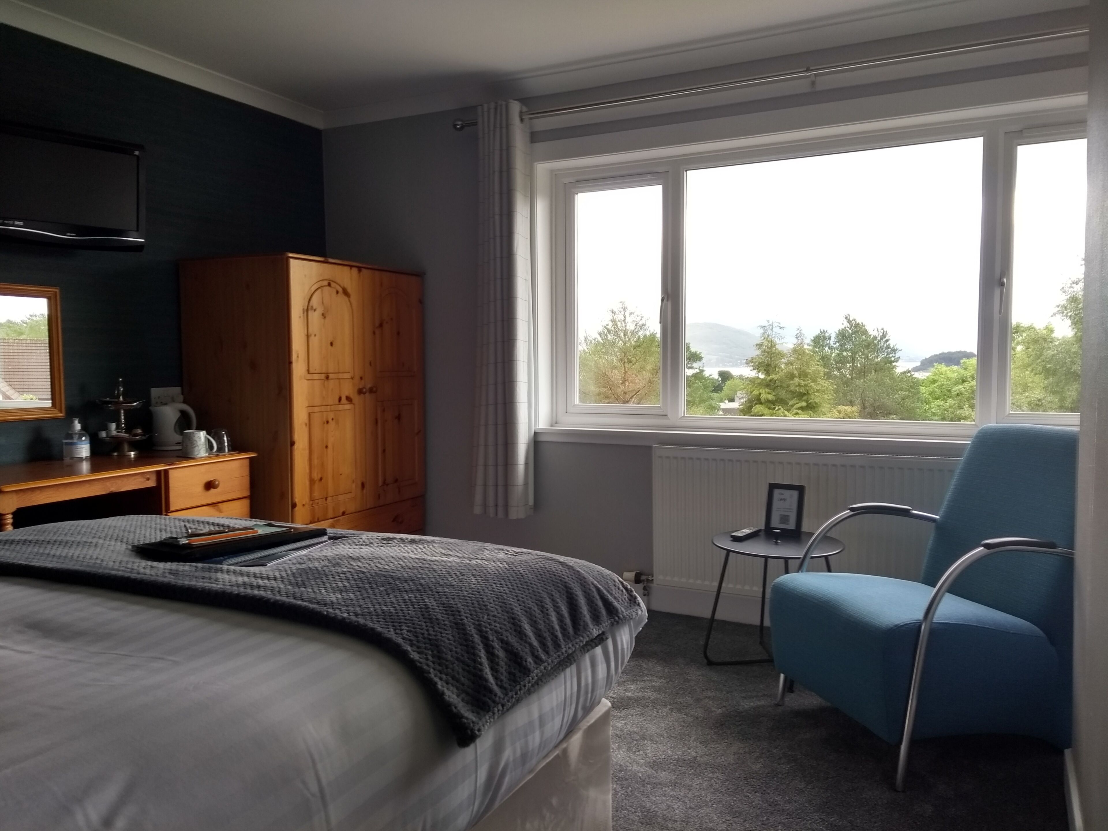 Een slaapkamer met een bed, een houten garderobe, een bureau met stoel, een raam met uitzicht op groen en heuvels, en een radiator.