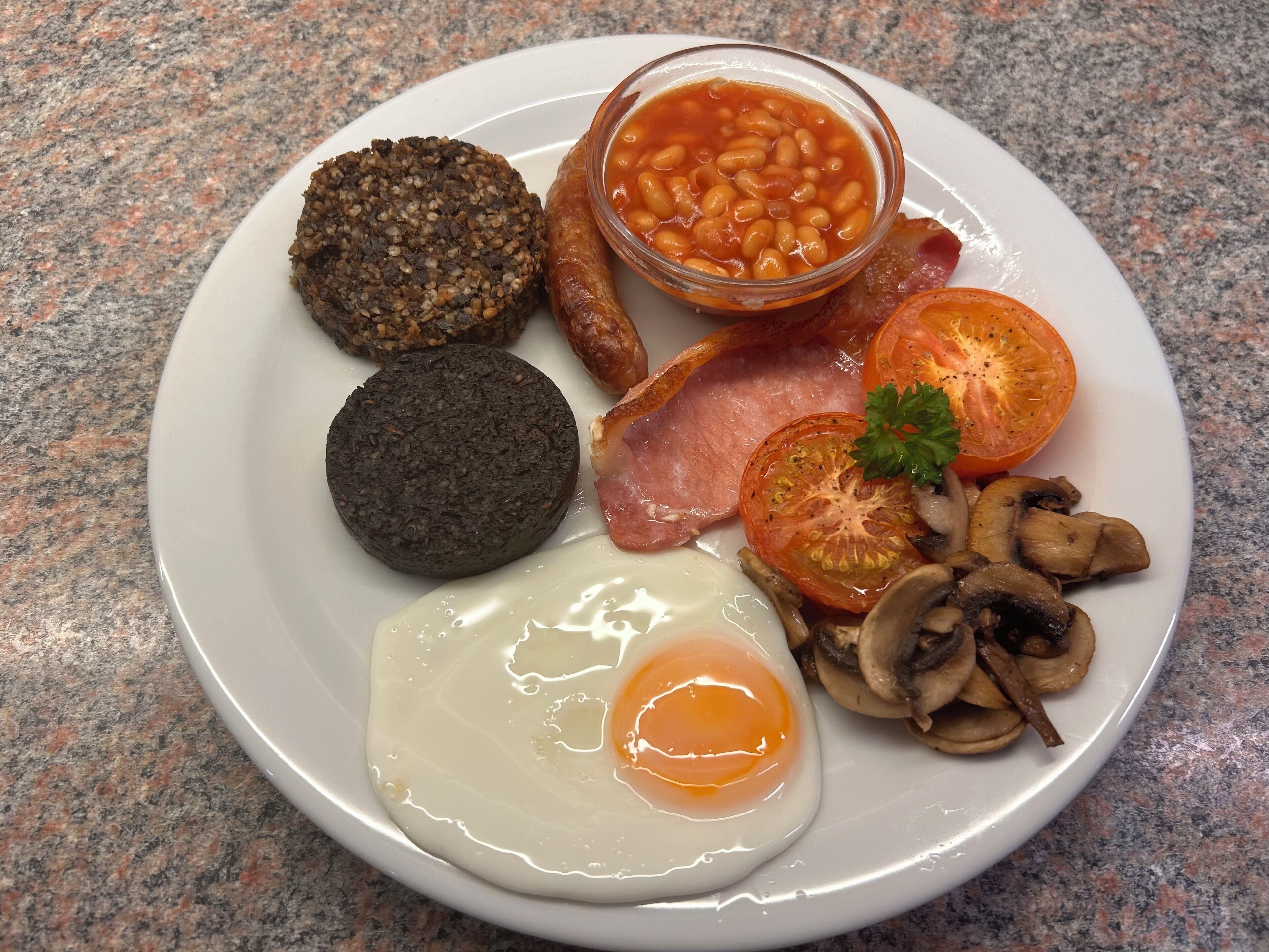 Une assiette de mets pour le petit-déjeuner comprenant des œufs, du bacon, des champignons, des tomates, des haricots et de la boudin noir.