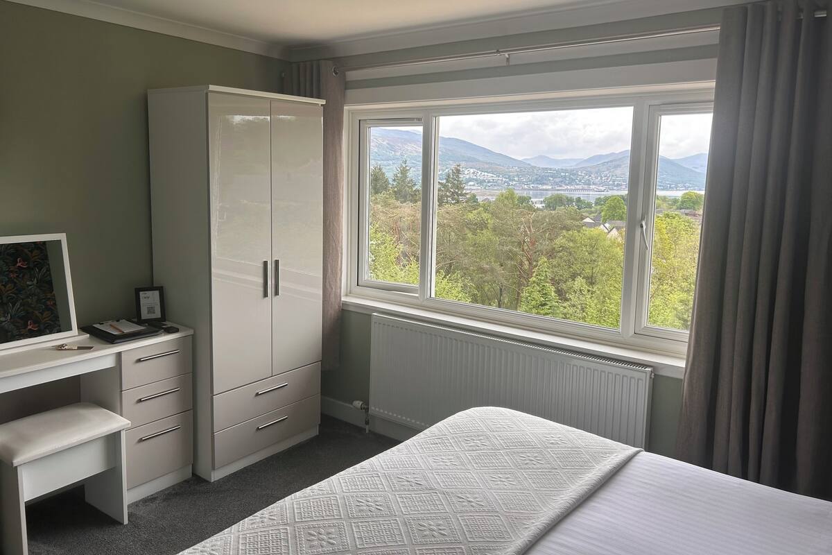 A bedroom with a bed, a desk, and a wardrobe. There is a window with a view of a landscape.