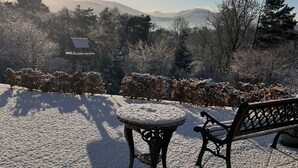 Schneebedeckte Landschaft mit einem Tisch und einer Bank, umgeben von Bäumen und einer entfernten Aussicht auf Berge.