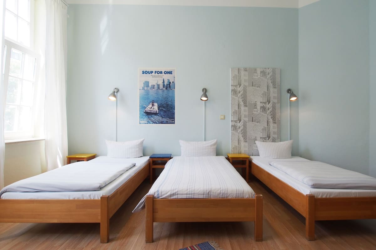 Quarto com três camas de solteiro, cada uma com estrutura de madeira e roupa de cama branca. Há duas mesas de cabeceira com abajures e um cartaz na parede.