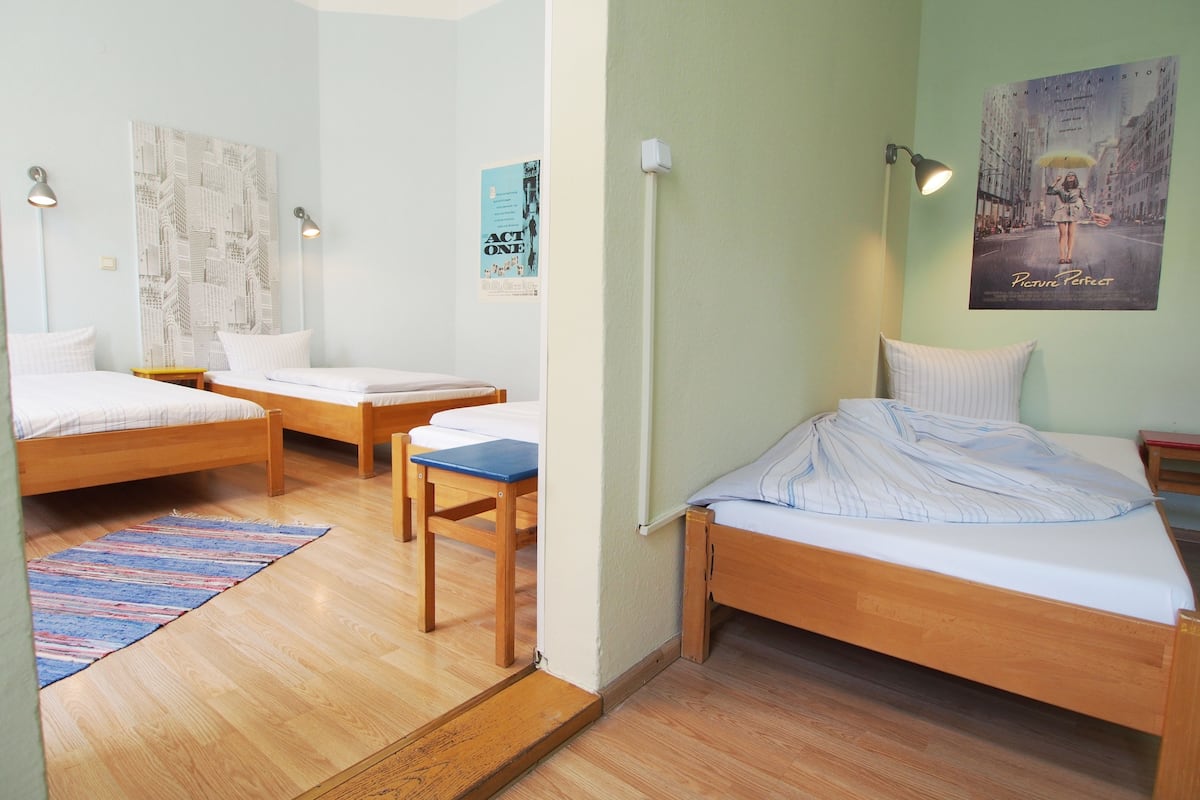 Quarto com três camas de madeira, uma mesa pequena e uma cadeira. Há cartazes nas paredes e um abajur no lado direito.