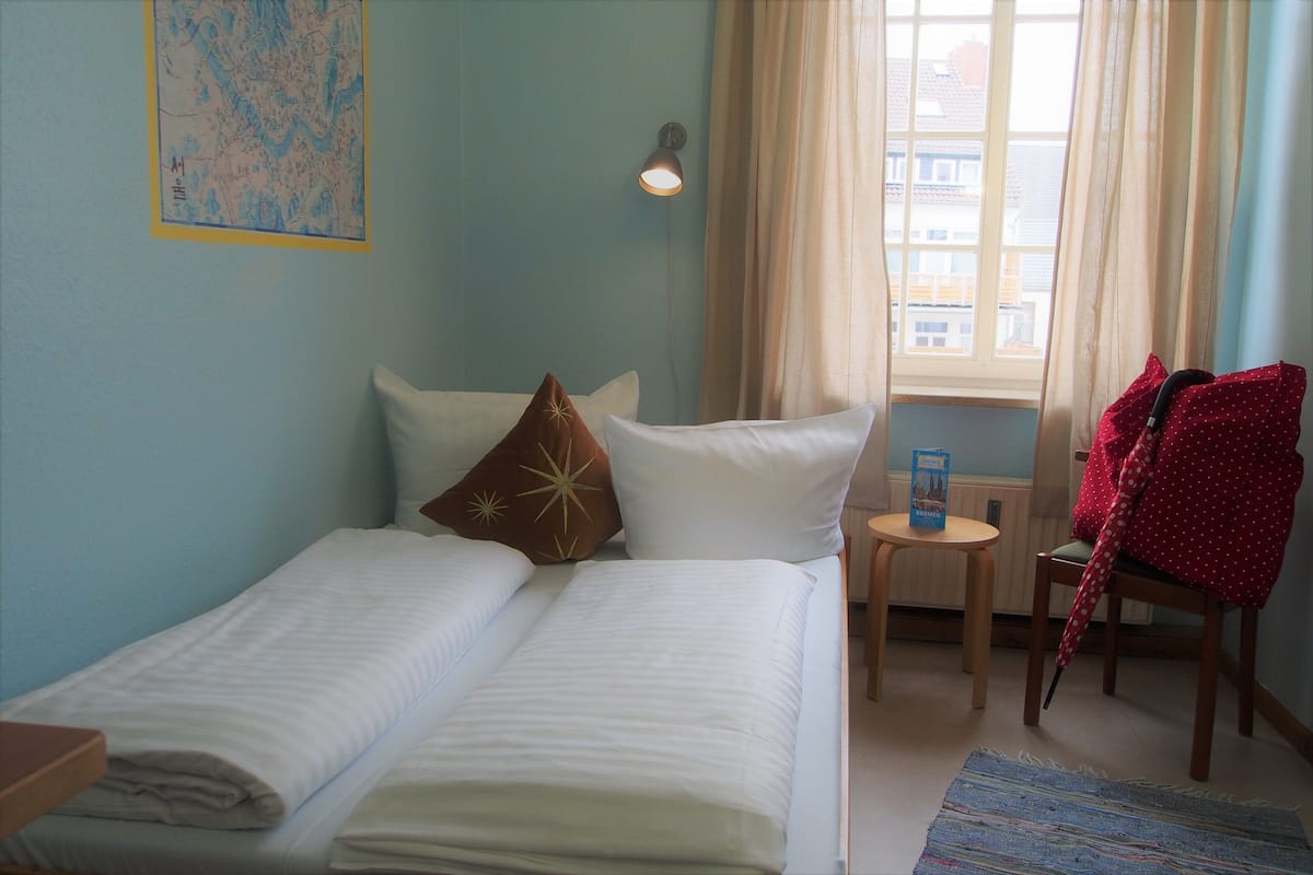 Uma cama de casal com roupa de cama branca, um mapa na parede, uma luminária e vista para prédios pela janela.