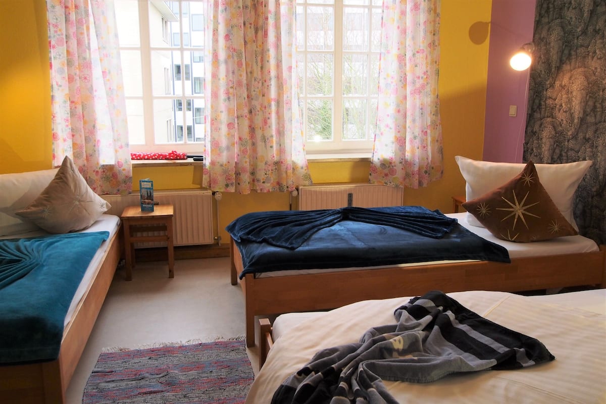 Quarto com duas camas, mesa de cabeceira, radiador e janela com cortinas.