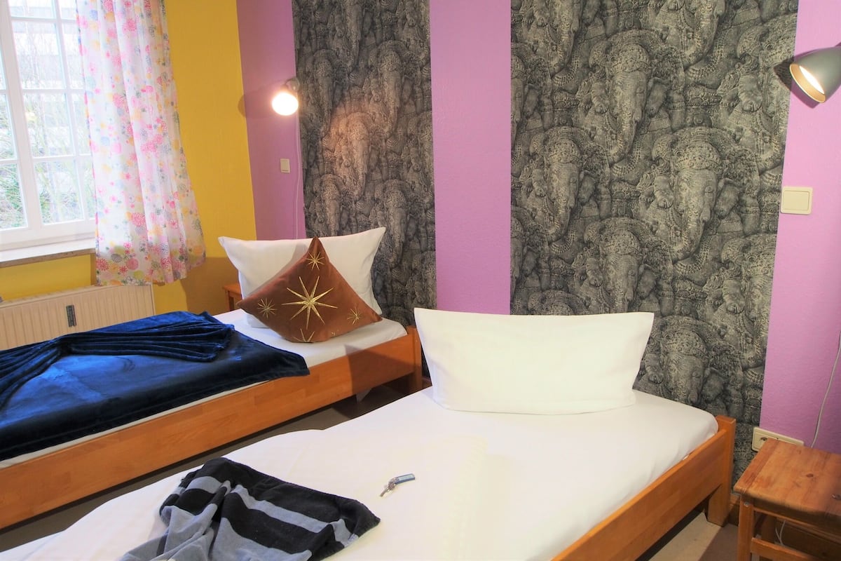 Quarto com duas camas, janela com cortinas florais e papel de parede estampado.